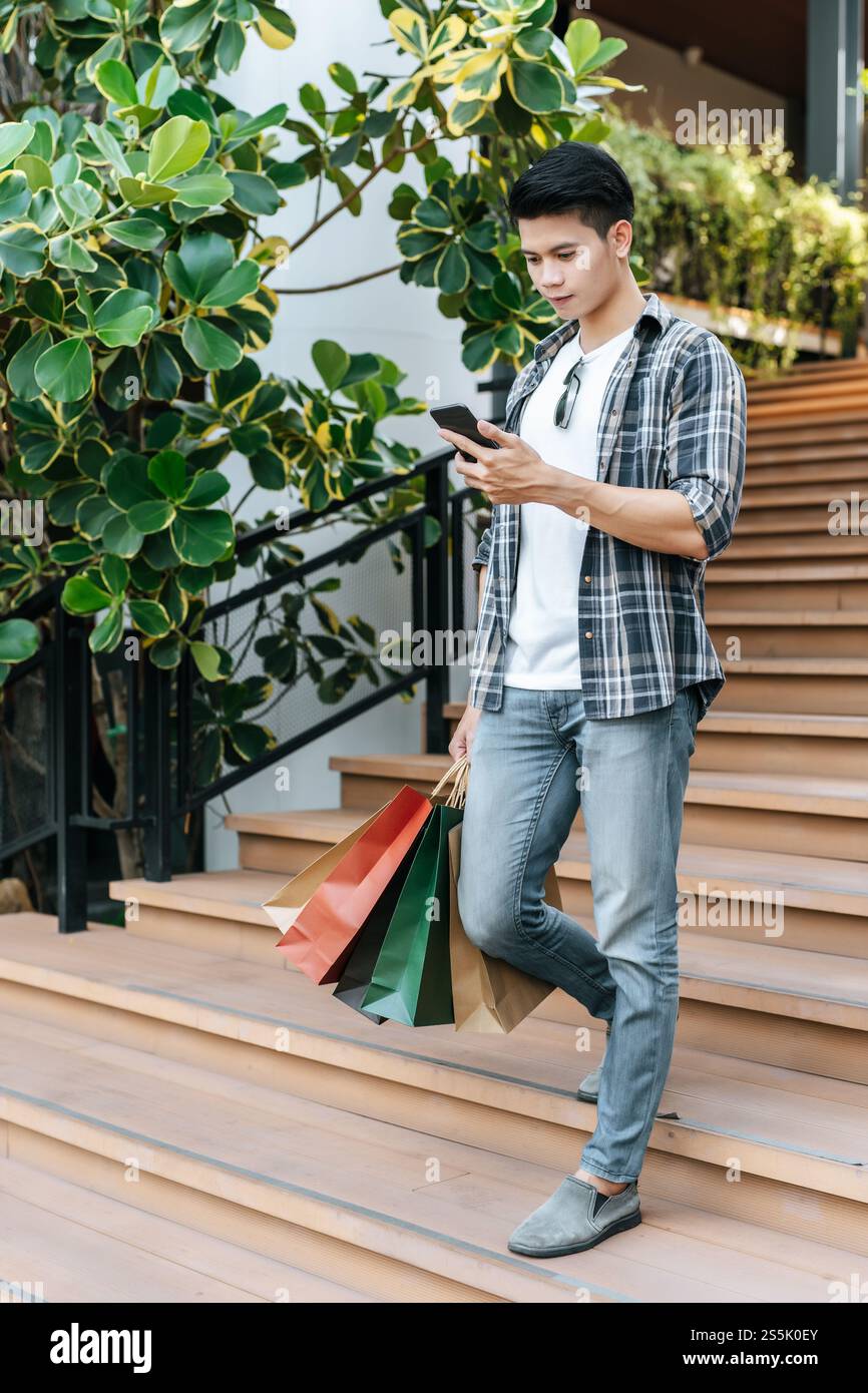 Portrait jeune homme beau tenant un sac en papier et utiliser un smartphone sur l'escalier du centre commercial, style de vie et nouveau concept normal, espace de copie Banque D'Images