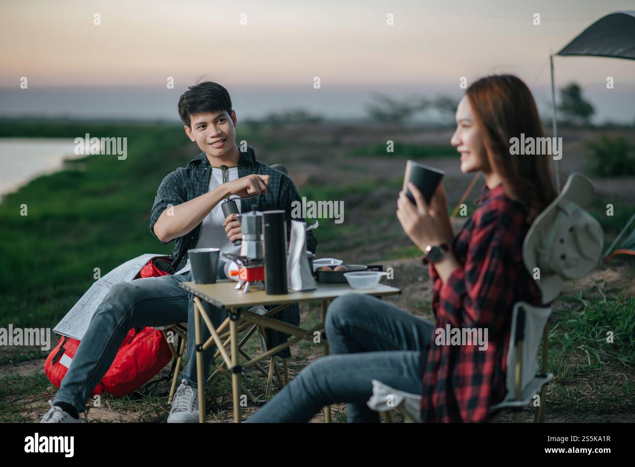 Mise au point sélective, bel homme faisant moulin à café frais, joyeux couple jeune routard assis devant la tente près du lac avec set de café Banque D'Images