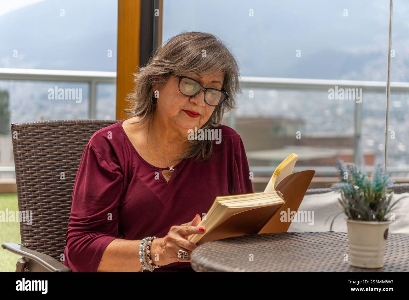 Portrait de femme âgée avec des lunettes et chemisier Bordeaux lisant un livre à une table en rotin à l'extérieur Banque D'Images