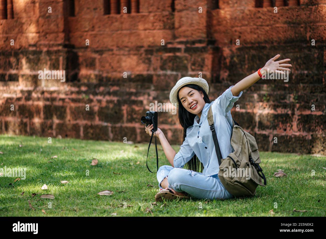 Jeune routard joyeux assis sur l'herbe levez son bras et souriant avec bonheur, tenant un appareil photo à la main tout en voyageant dans un site antique Banque D'Images