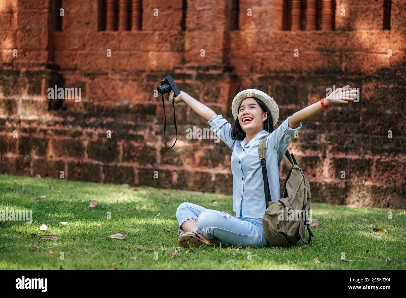 Jeune routard joyeux assis sur l'herbe levez son bras et souriant avec bonheur, tenant un appareil photo à la main tout en voyageant dans un site antique Banque D'Images