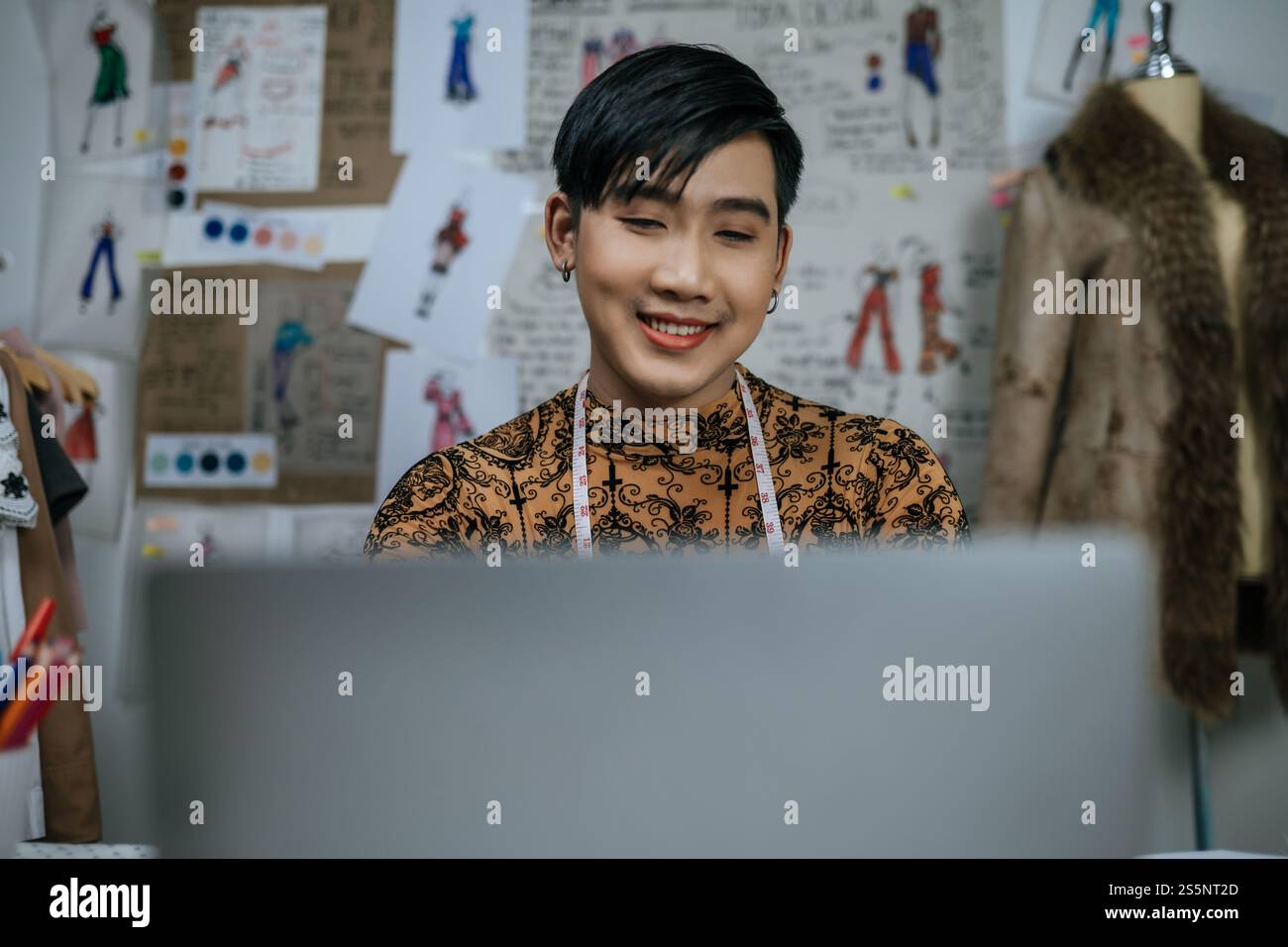 Gros plan visage de jeune tailleur masculin asiatique professionnel avec ruban à mesurer sur le cou travaillant sur ordinateur portable avec fond de dessins en studio. Concepteur et Banque D'Images