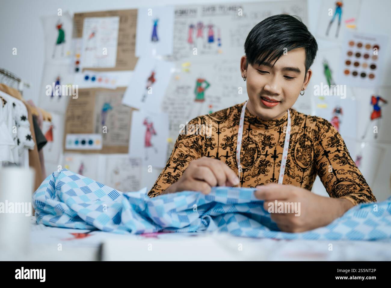 Portrait de jeune tailleur masculin asiatique heureux professionnel avec ruban à mesurer sur le cou travaillant avec des dessins de fond en studio. Designer et LGBTQ Banque D'Images