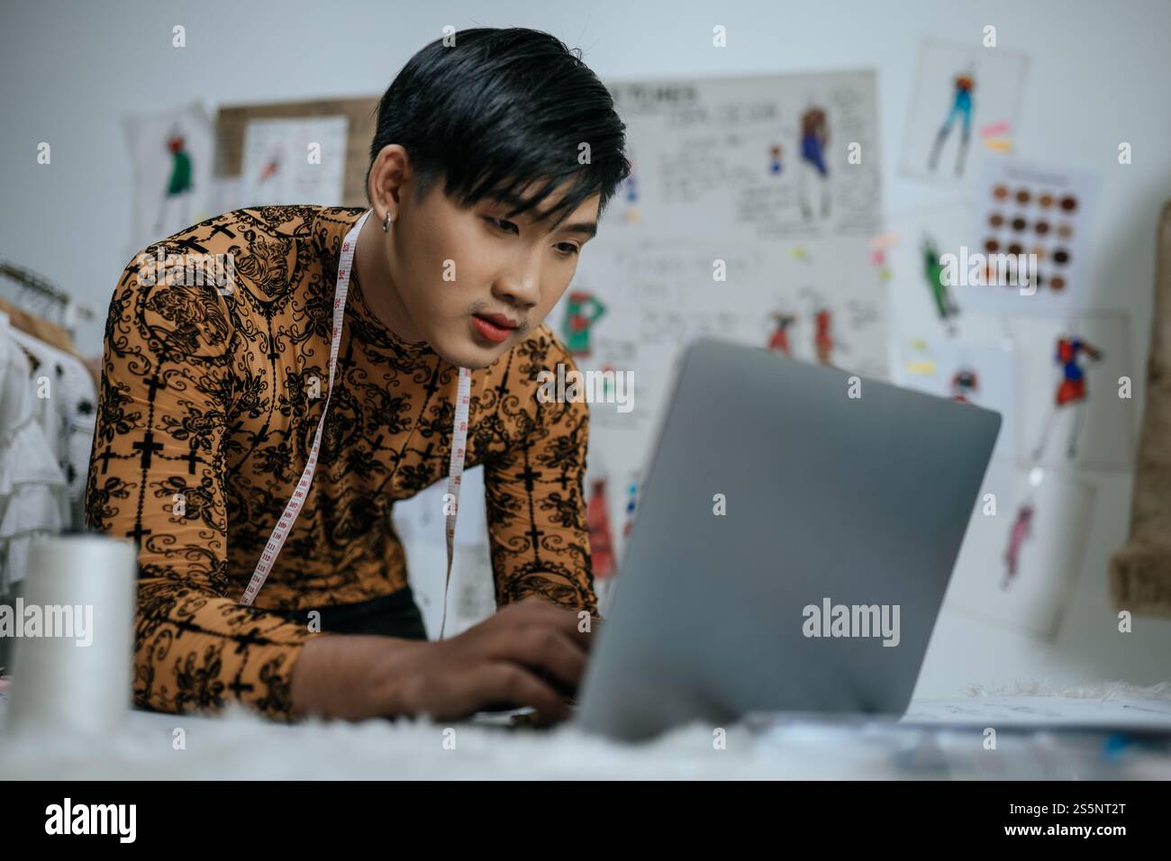 Portrait de jeune tailleur masculin asiatique professionnel avec ruban à mesurer sur le cou travaillant sur ordinateur portable avec fond de dessins en studio. Designer et LGBTQ Banque D'Images