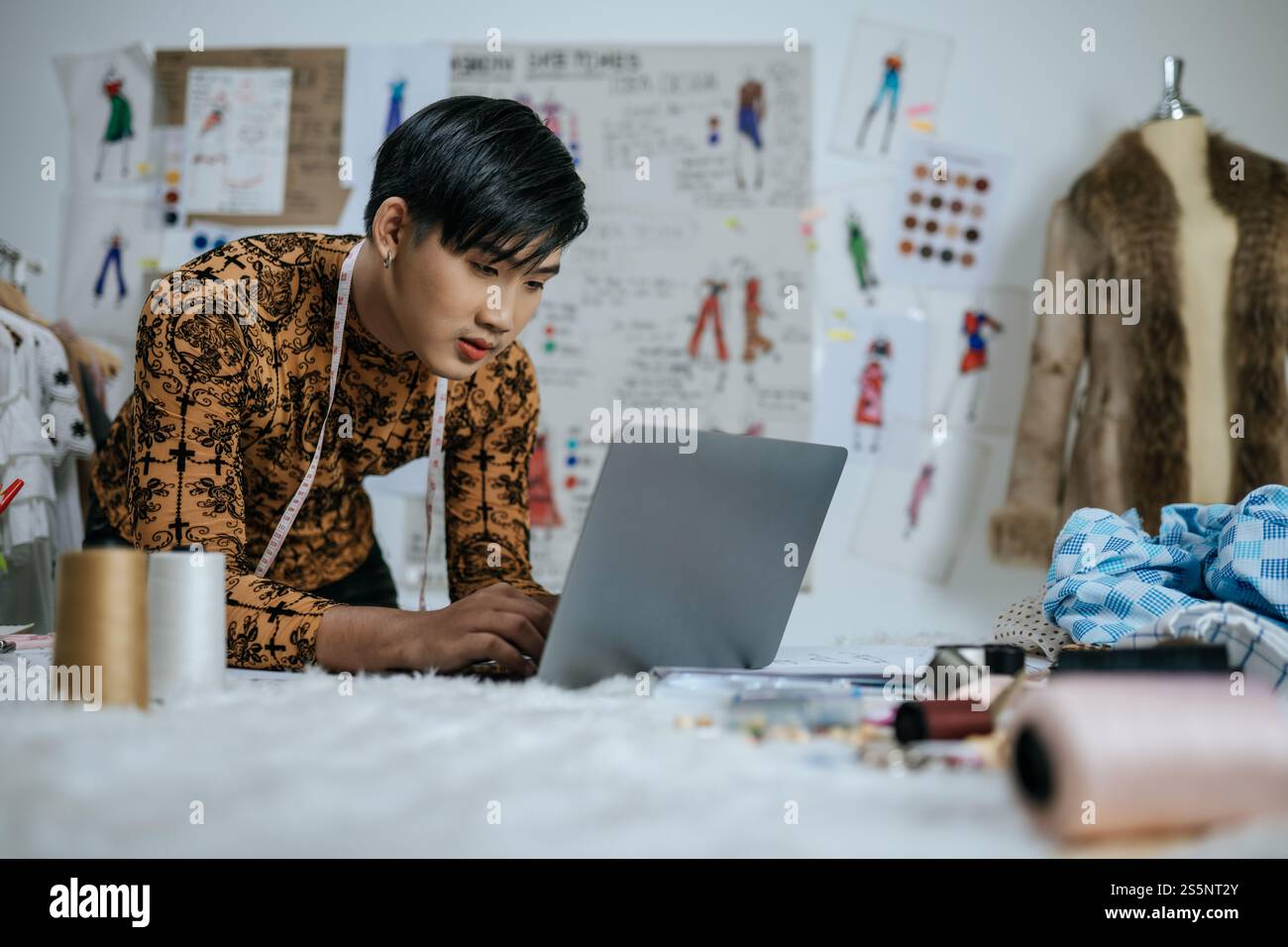 Portrait de jeune tailleur masculin asiatique professionnel avec ruban à mesurer sur le cou travaillant sur ordinateur portable avec fond de dessins en studio. Designer et LGBTQ Banque D'Images