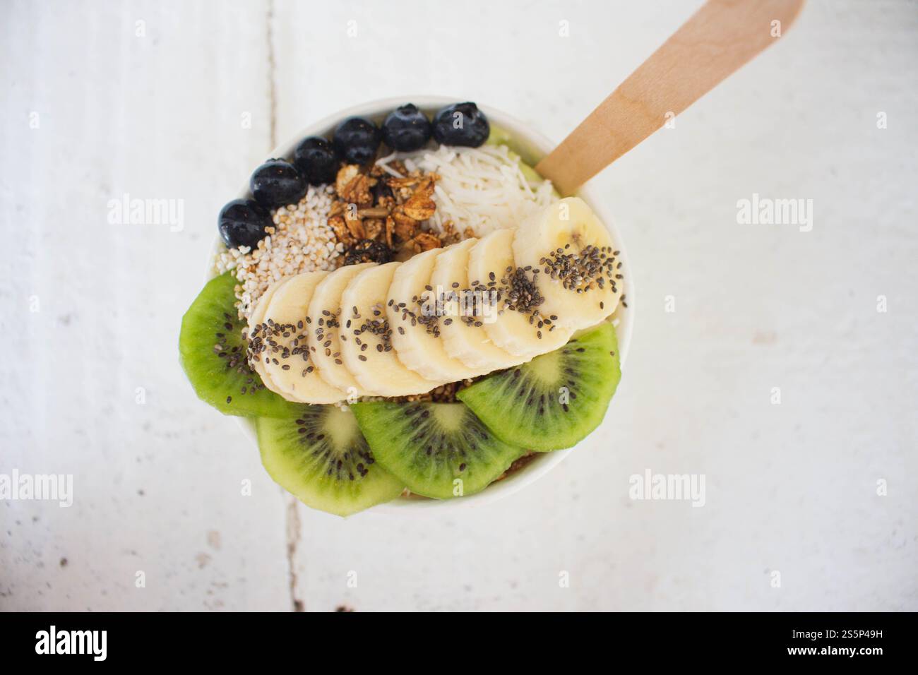 Un bol d'açai avec des myrtilles, des graines de chia, des kiwis, du granola et de la noix de coco râpée est sur une table en bois blanc avec une cuillère en bois Banque D'Images