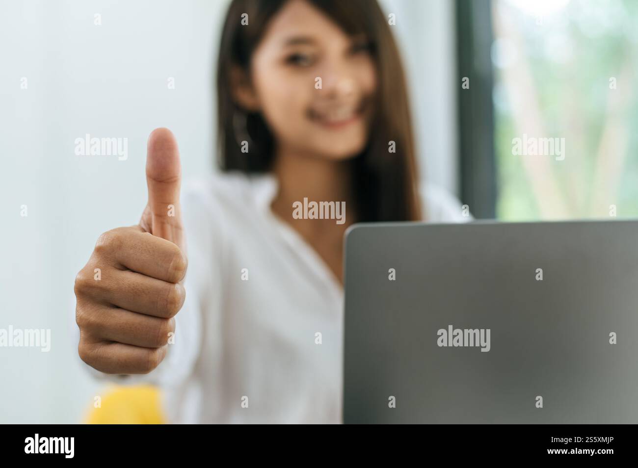 Mise au point sélective sur la main féminine avec le pouce vers le haut, brouillé asiatique beau sourire de femme avec heureux pendant le travail sur l'ordinateur portable au café Banque D'Images