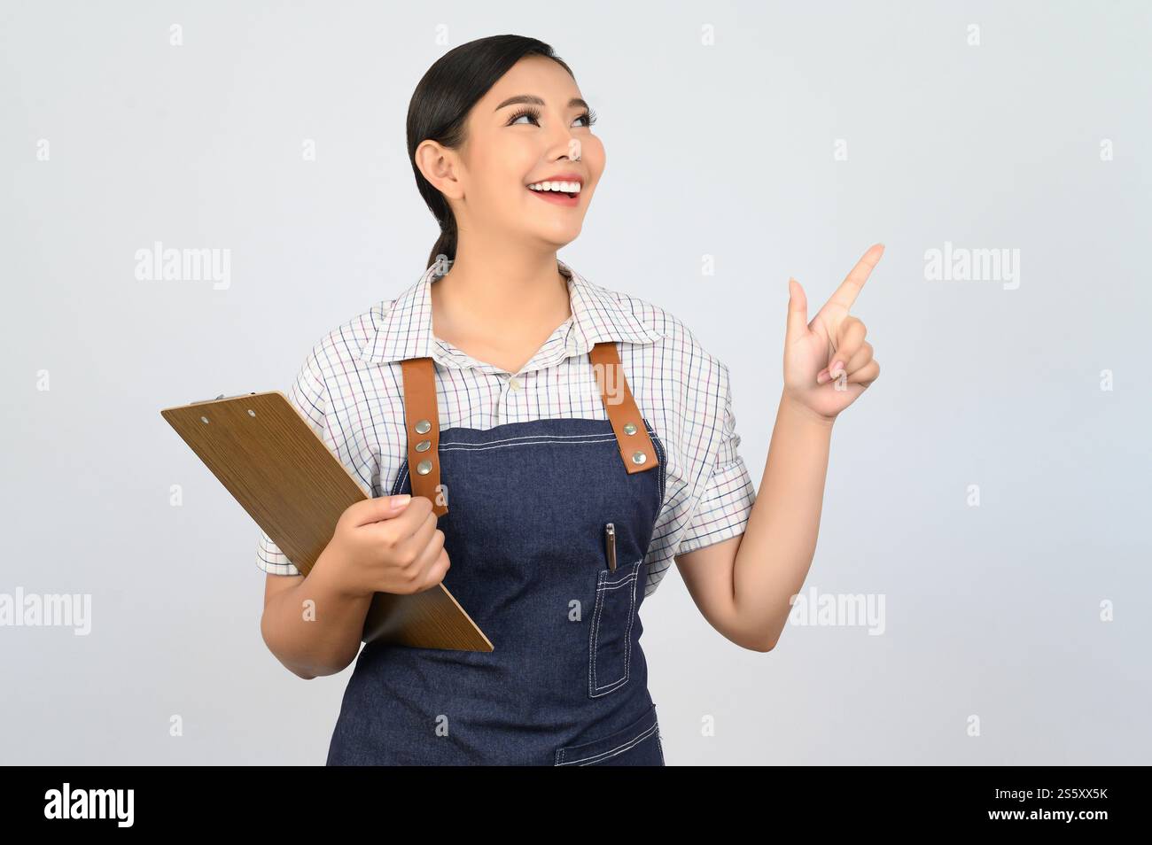 Portrait de jeune femme asiatique dans l'uniforme de serveuse tenant le presse-papiers et le doigt de pointe, copiez l'espace pour insérer des produits pour la publicité isolé sur Banque D'Images