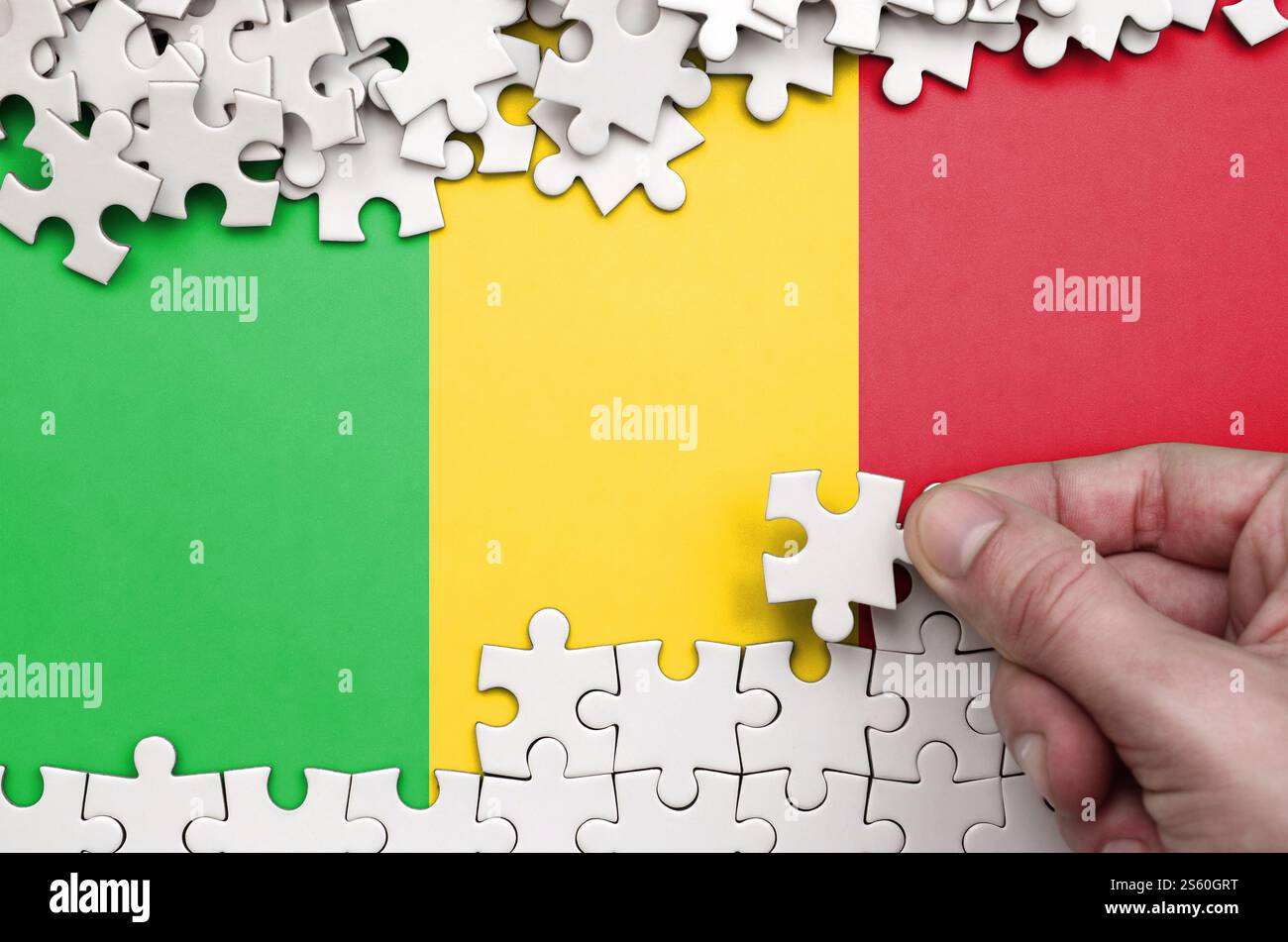 Le drapeau du Mali est représenté sur une table sur laquelle la main humaine plie un puzzle de couleur blanche. Le drapeau du Mali est représenté sur une table sur laquelle la main humaine Banque D'Images