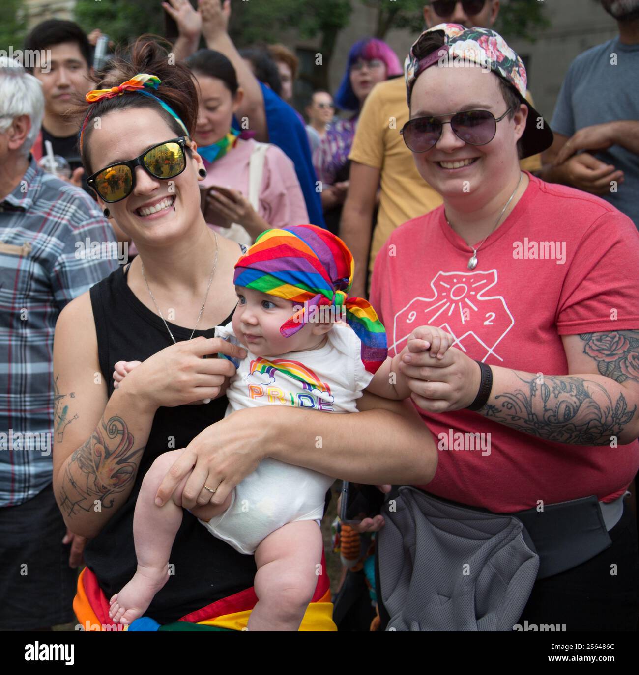 Une famille LGBTQ+ heureuse sourit tout en célébrant l’événement fierté de Montréal. Le couple tient son bébé, qui porte un foulard arc-en-ciel et un « PRI Banque D'Images