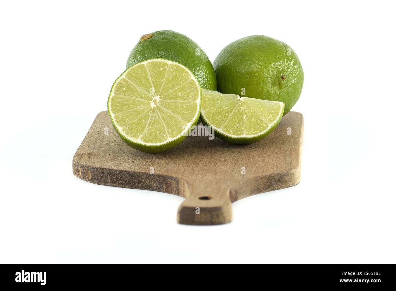 Avec deux citrons verts entiers à côté d'un citron vert tranché et d'un coin de citron vert, le tout disposé sur une petite planche à découper en bois sur un fond blanc propre Banque D'Images