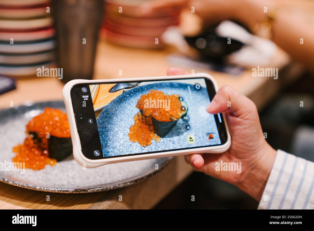 Une main tenant un smartphone, prenant un gros plan d'un plat de sushis magnifiquement présenté surmonté d'œufs de saumon sur une assiette élégante Banque D'Images