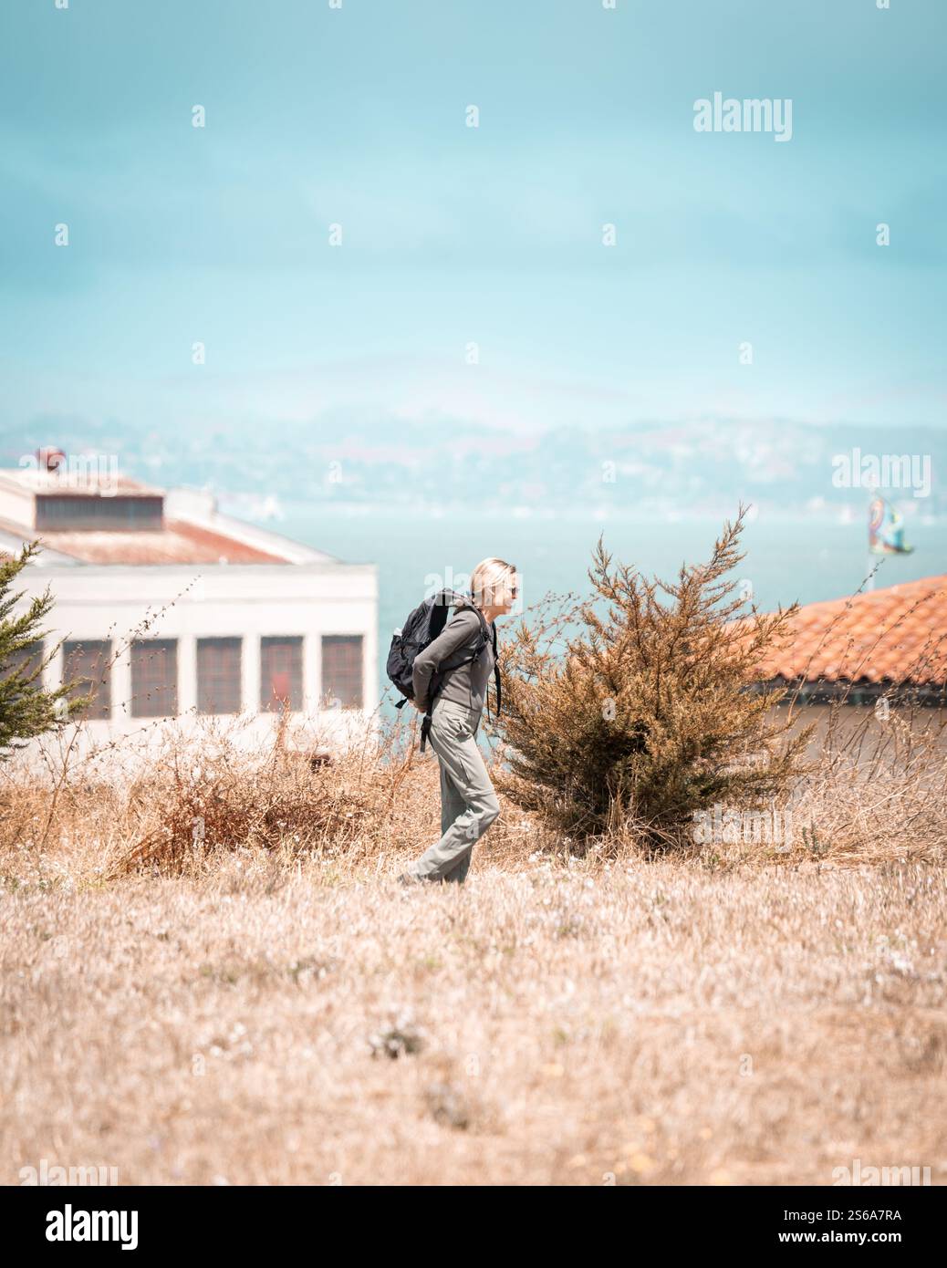 Une femme marchant dans des champs d'herbe sèche près des bâtiments côtiers par une journée ensoleillée, portant un sac à dos et une tenue verte, avec un ciel bleu vibrant Banque D'Images