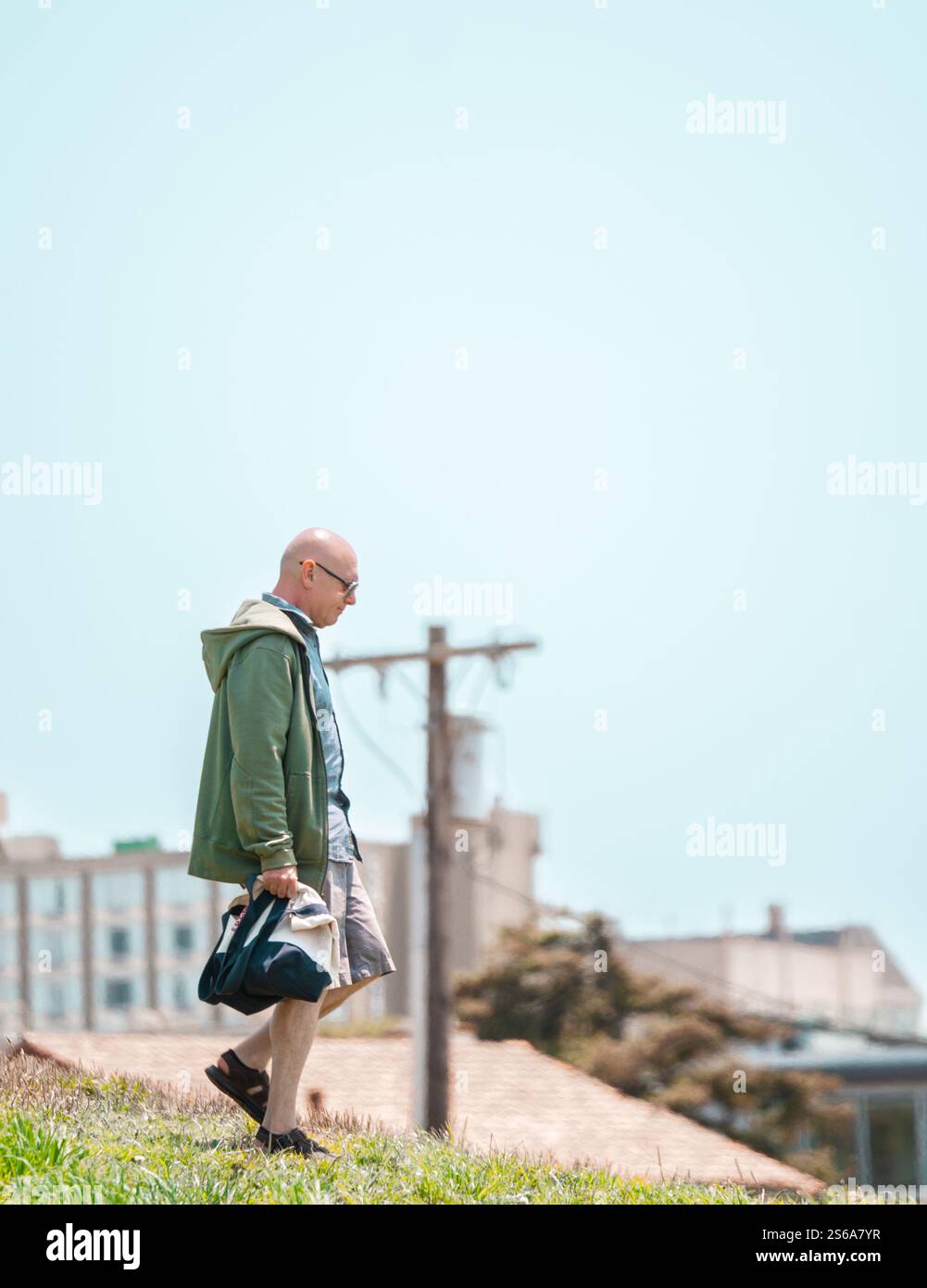 Un homme marchant sur une pente herbeuse, portant un sac, avec un paysage urbain et un poteau utilitaire en arrière-plan, sous un ciel bleu clair par une journée ensoleillée Banque D'Images