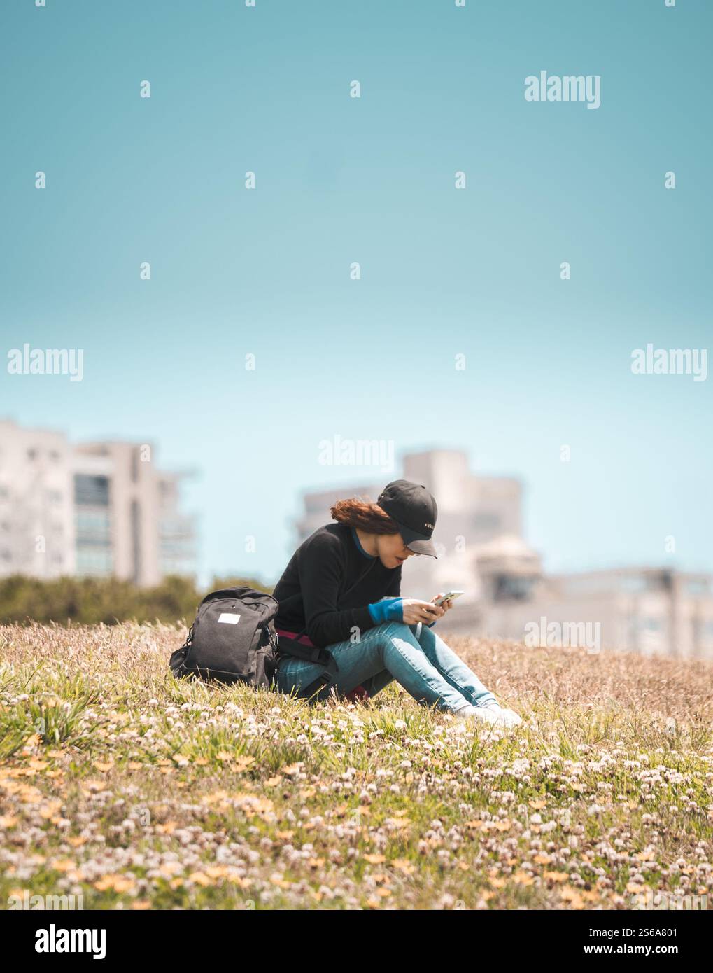 Une jeune femme assise sur un champ herbeux entouré de fleurs sauvages, englouti dans son téléphone, avec un sac à dos à ses côtés Banque D'Images