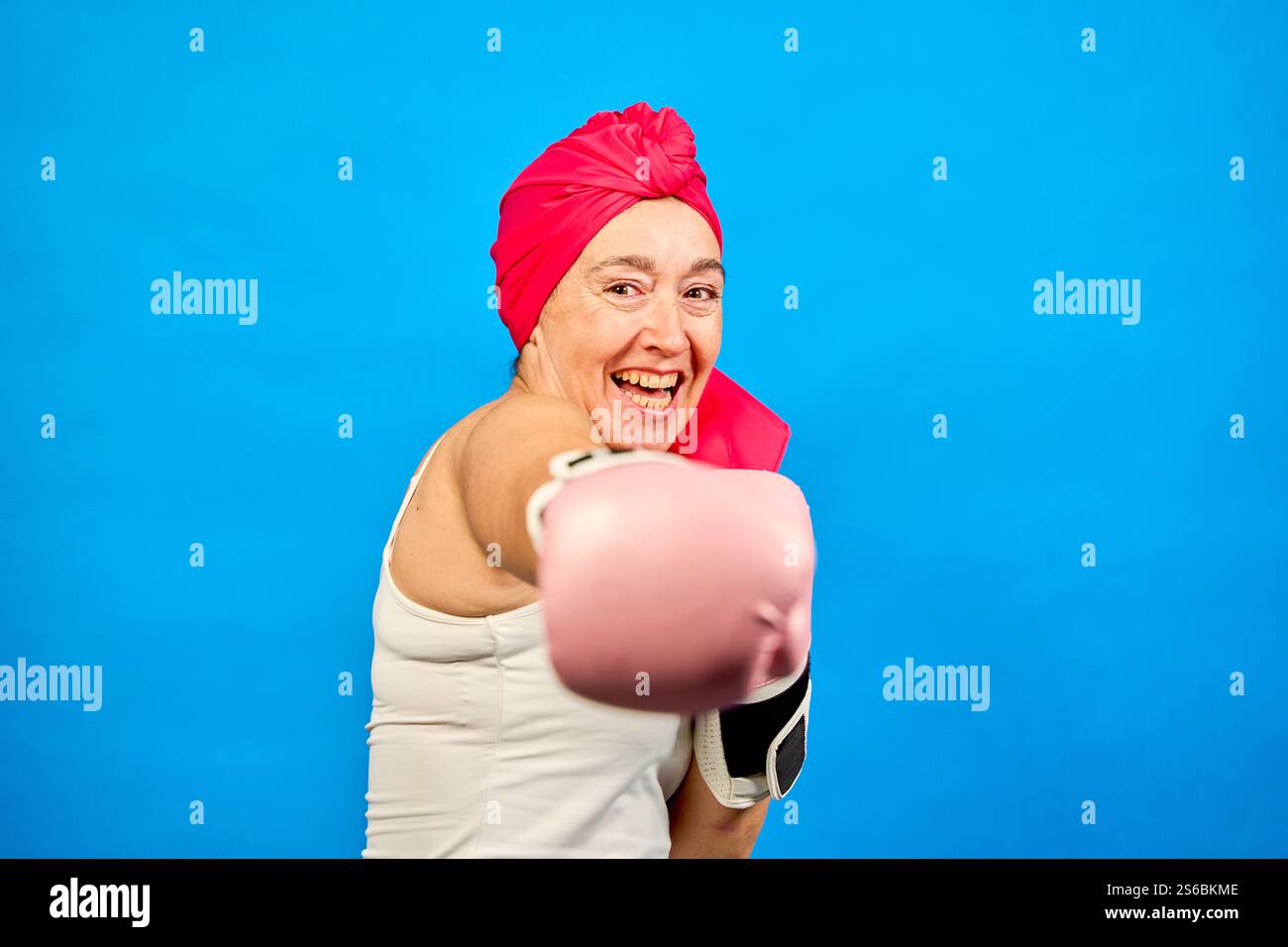 Studio tourné sur fond bleu d'une femme d'âge moyen souriante portant des gants de boxe roses et un foulard, frappant et combattant le cancer Banque D'Images