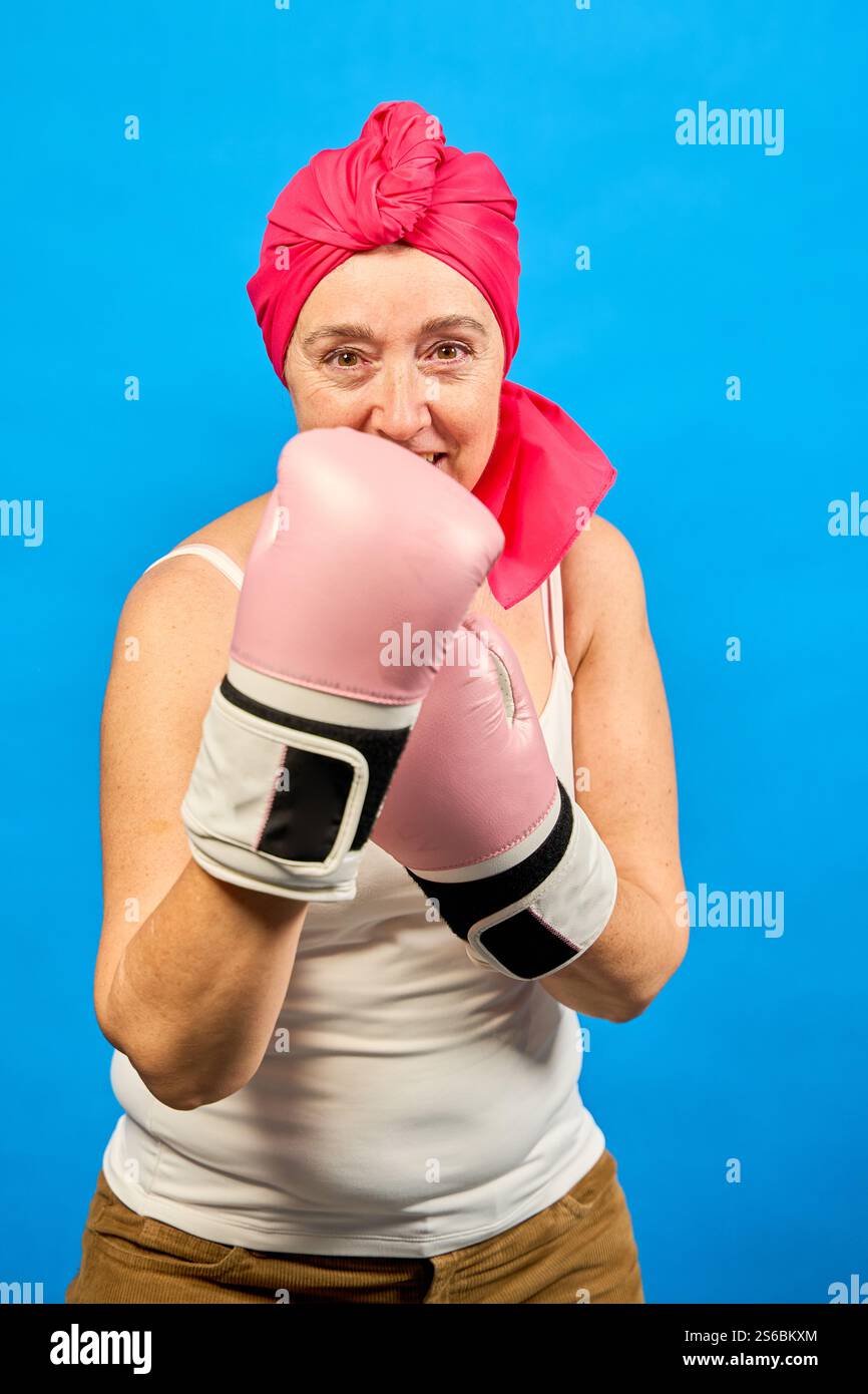 Femme mature souriante portant des gants de boxe roses et foulard combattant le cancer avec une attitude positive Banque D'Images