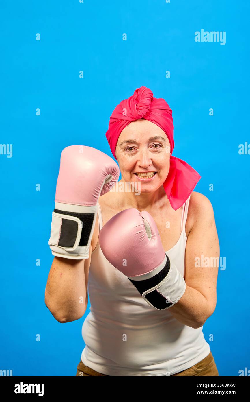 Femme d'âge moyen portant des gants de boxe roses et un foulard luttant contre le cancer avec une expression déterminée sur fond bleu Banque D'Images