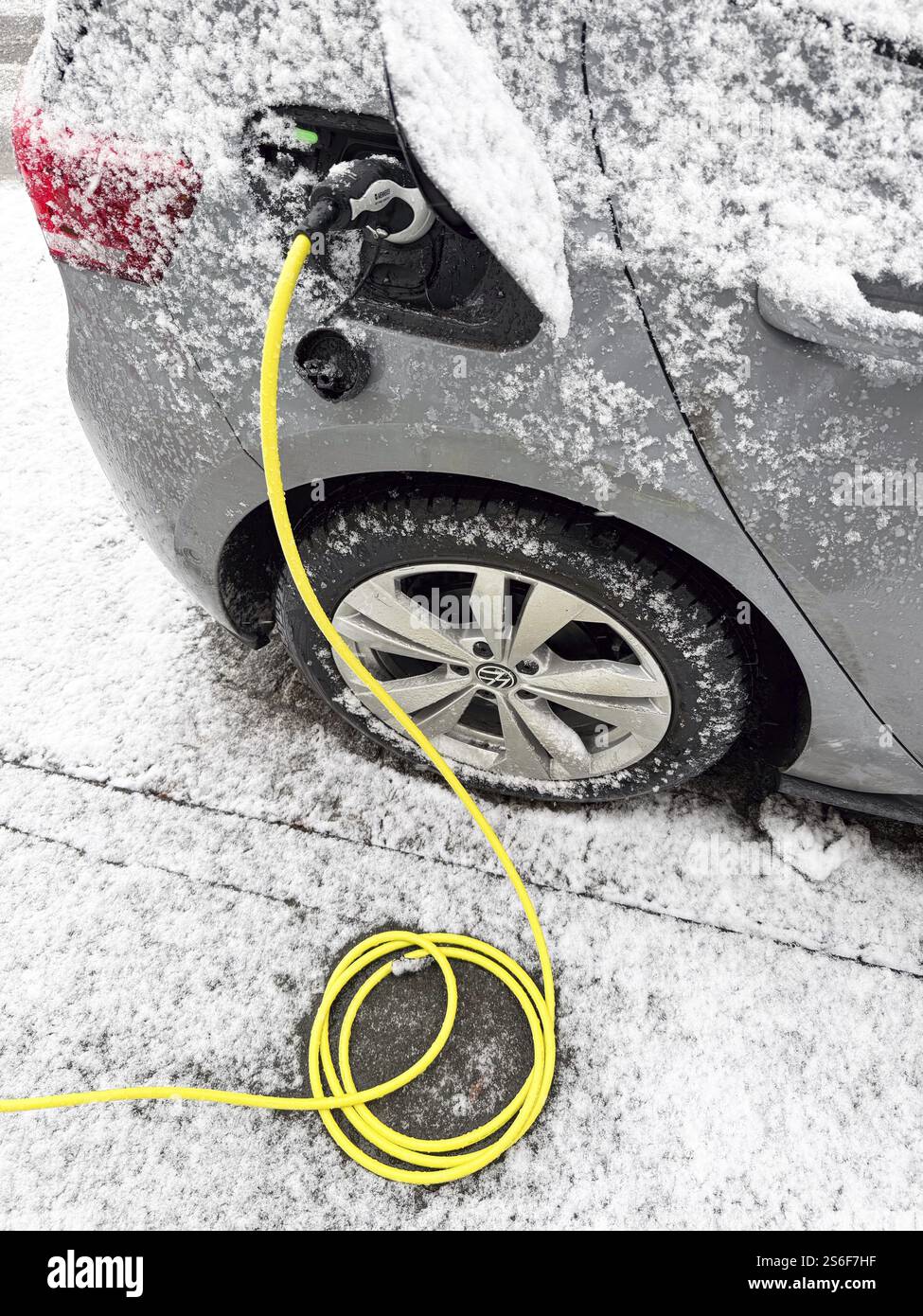 Recharge d'un véhicule électrique en hiver. Un câble de charge jaune est branché sur la prise de charge de la voiture alors que de la neige s'est accumulée sur la carrosserie, Banque D'Images