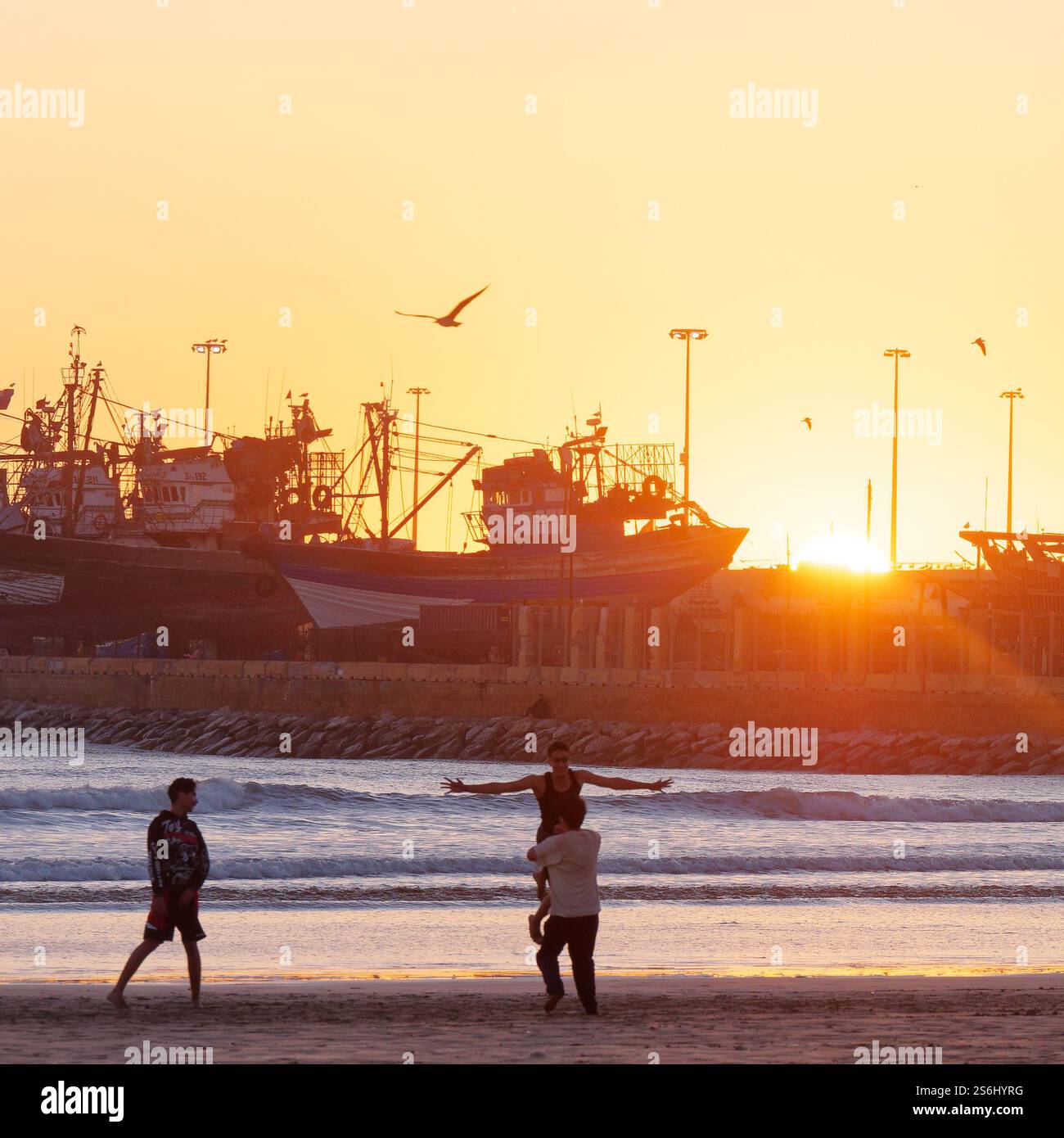 Les jeunes hommes jouent sur la plage tandis que l'un soulève l'autre au coucher du soleil avec le chantier naval derrière à Essaouira, au Maroc. Jan 2025 Banque D'Images