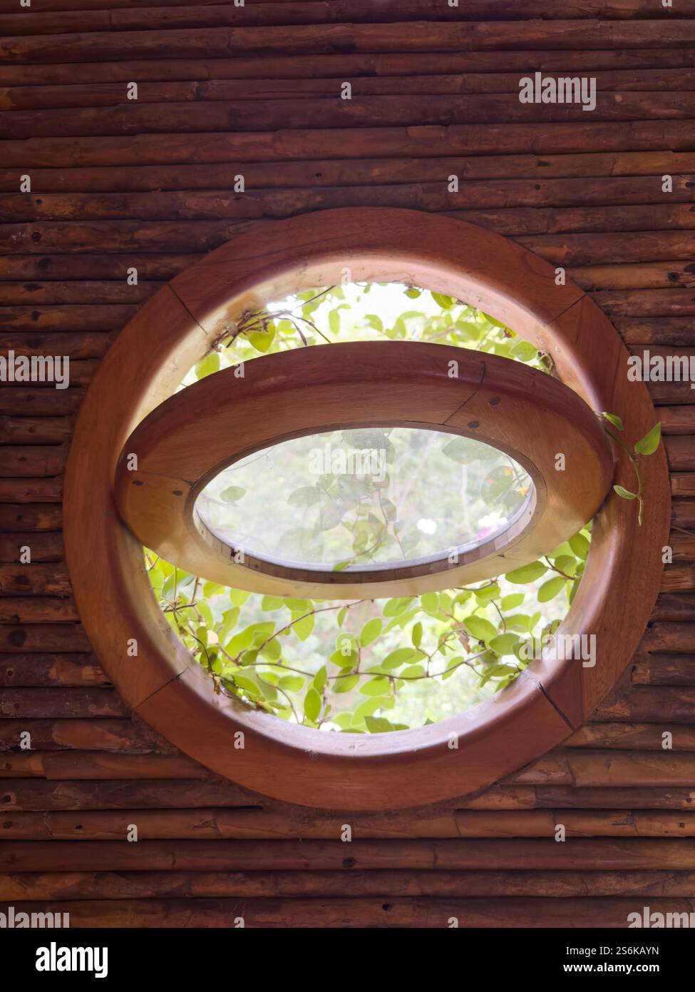 La lumière naturelle filtre à travers une fenêtre circulaire en bois entourée de verdure et de mur rustique, créant une atmosphère sereine. Banque D'Images