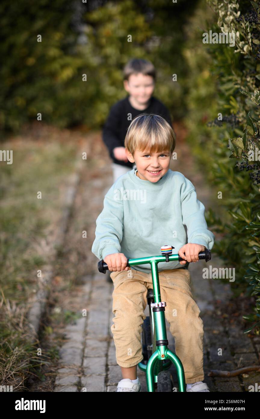 Garçons chevauchant dans un jardin le long d'un chemin pavé, l'un sur un vélo d'équilibre et l'autre sur un scooter, entouré de verdure et de lumière du soleil. Banque D'Images