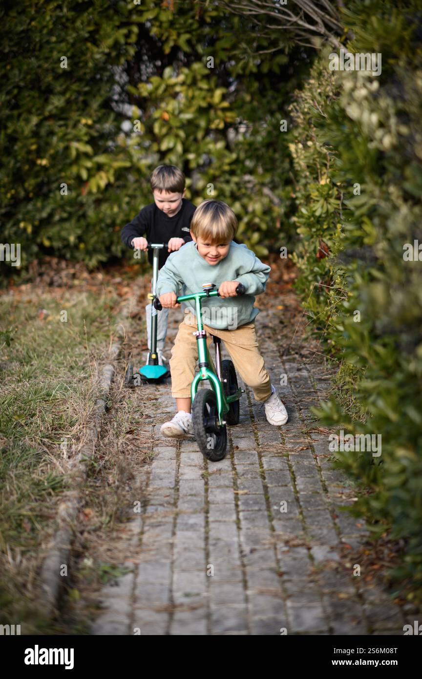 Garçons chevauchant dans un jardin le long d'un chemin pavé, l'un sur un vélo d'équilibre et l'autre sur un scooter, entouré de verdure et de lumière du soleil. Banque D'Images