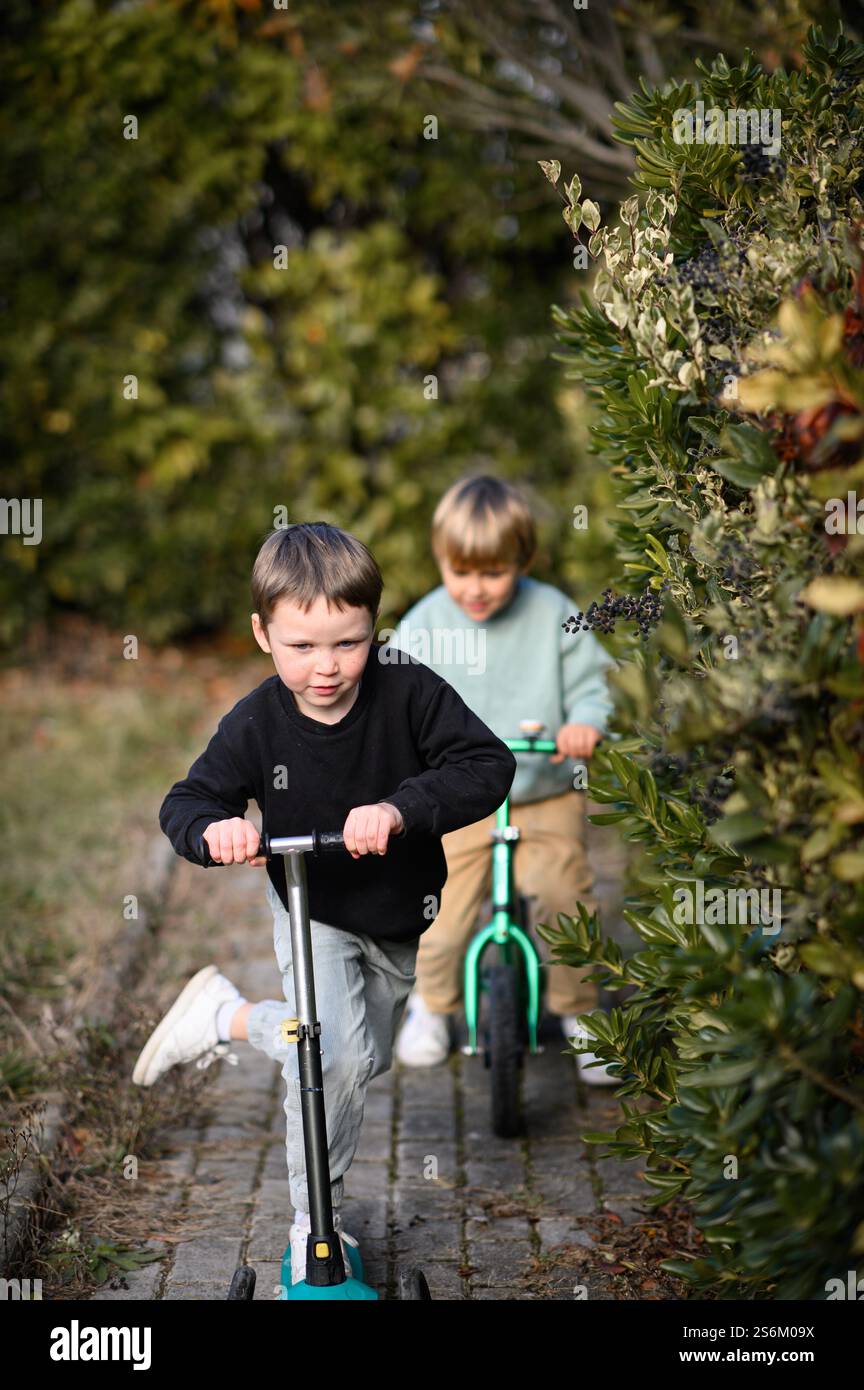 Garçons chevauchant dans un jardin le long d'un chemin pavé, l'un sur un vélo d'équilibre et l'autre sur un scooter, entouré de verdure et de lumière du soleil. Banque D'Images