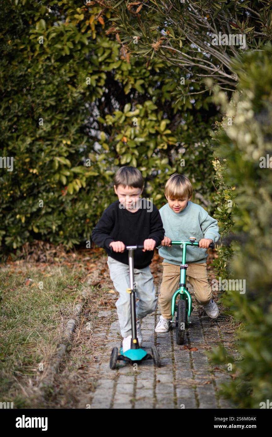 Garçons chevauchant dans un jardin le long d'un chemin pavé, l'un sur un vélo d'équilibre et l'autre sur un scooter, entouré de verdure et de lumière du soleil. Banque D'Images