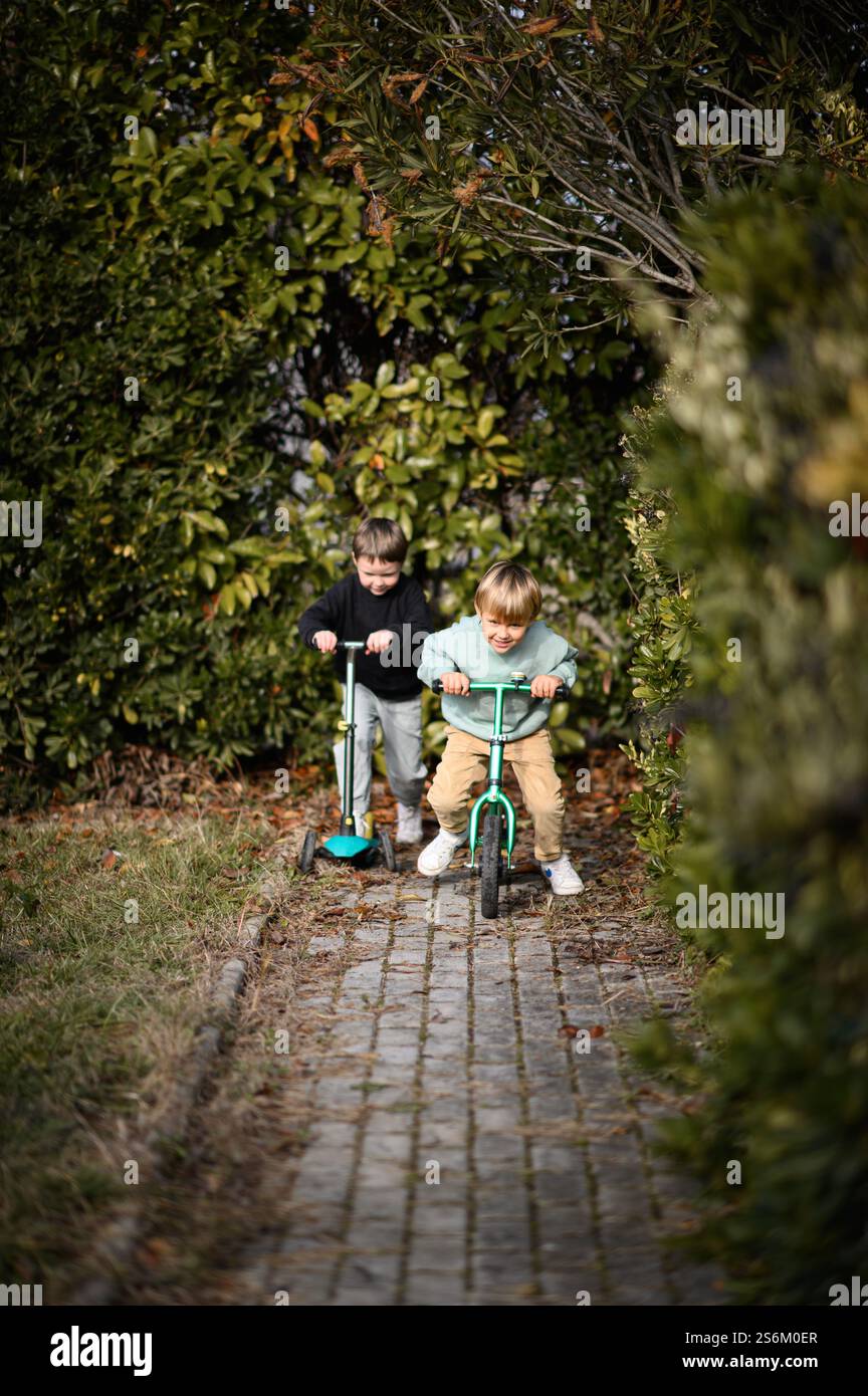 Garçons chevauchant dans un jardin le long d'un chemin pavé, l'un sur un vélo d'équilibre et l'autre sur un scooter, entouré de verdure et de lumière du soleil. Banque D'Images