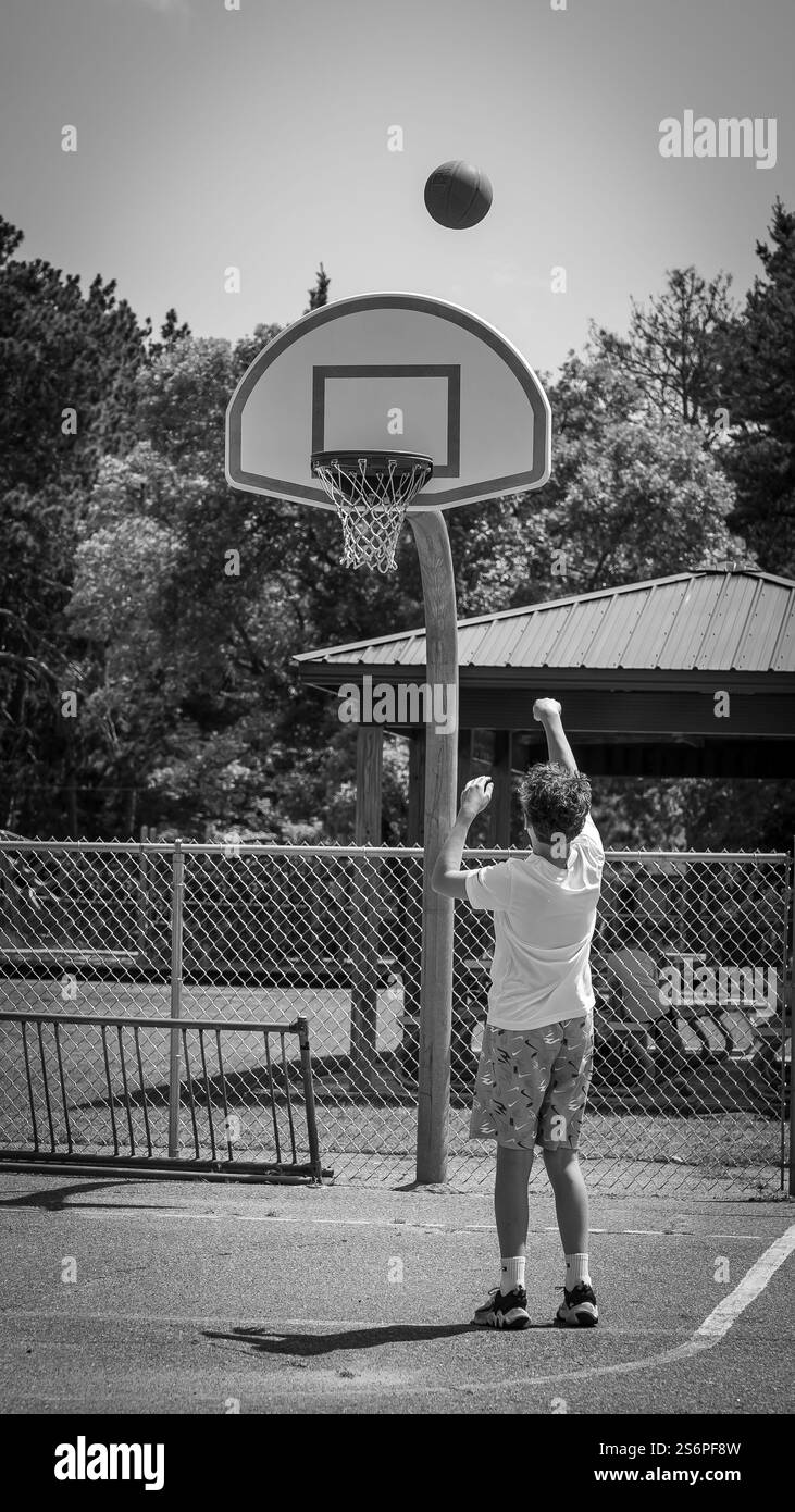 Un jeune athlète jouant au basket-ball sur un terrain de quartier, tirant la balle sur le panier, sur une photo en noir et blanc. Banque D'Images