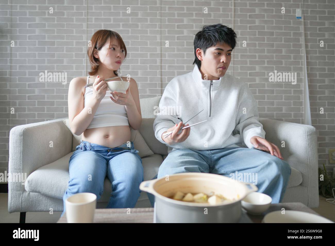 Une femme japonaise enceinte d'une vingtaine d'années et son mari, habillés pour montrer leur estomac, s'assoient sur un canapé dans un appartement. Banque D'Images
