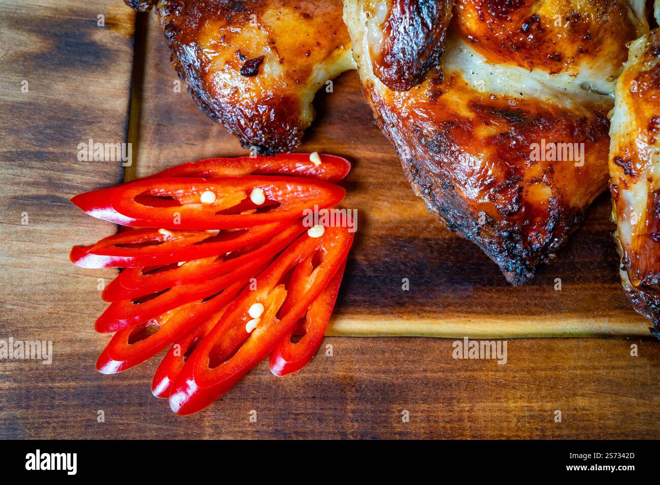 Le poulet parfaitement grillé repose à côté de tranches vibrantes de piment rouge sur une planche de bois rustique, présentant une présentation culinaire attrayante idéale pour un Banque D'Images