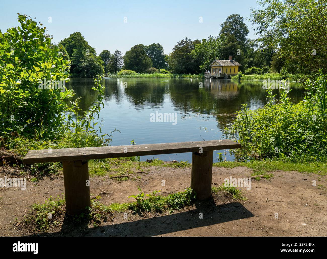 Banc de parc en bois surplombant le lac Belton House et le hangar à bateaux lors d'une journée d'été ensoleillée en juin avec un ciel bleu clair, Grantham, Lincolnshire, Angleterre. Banque D'Images
