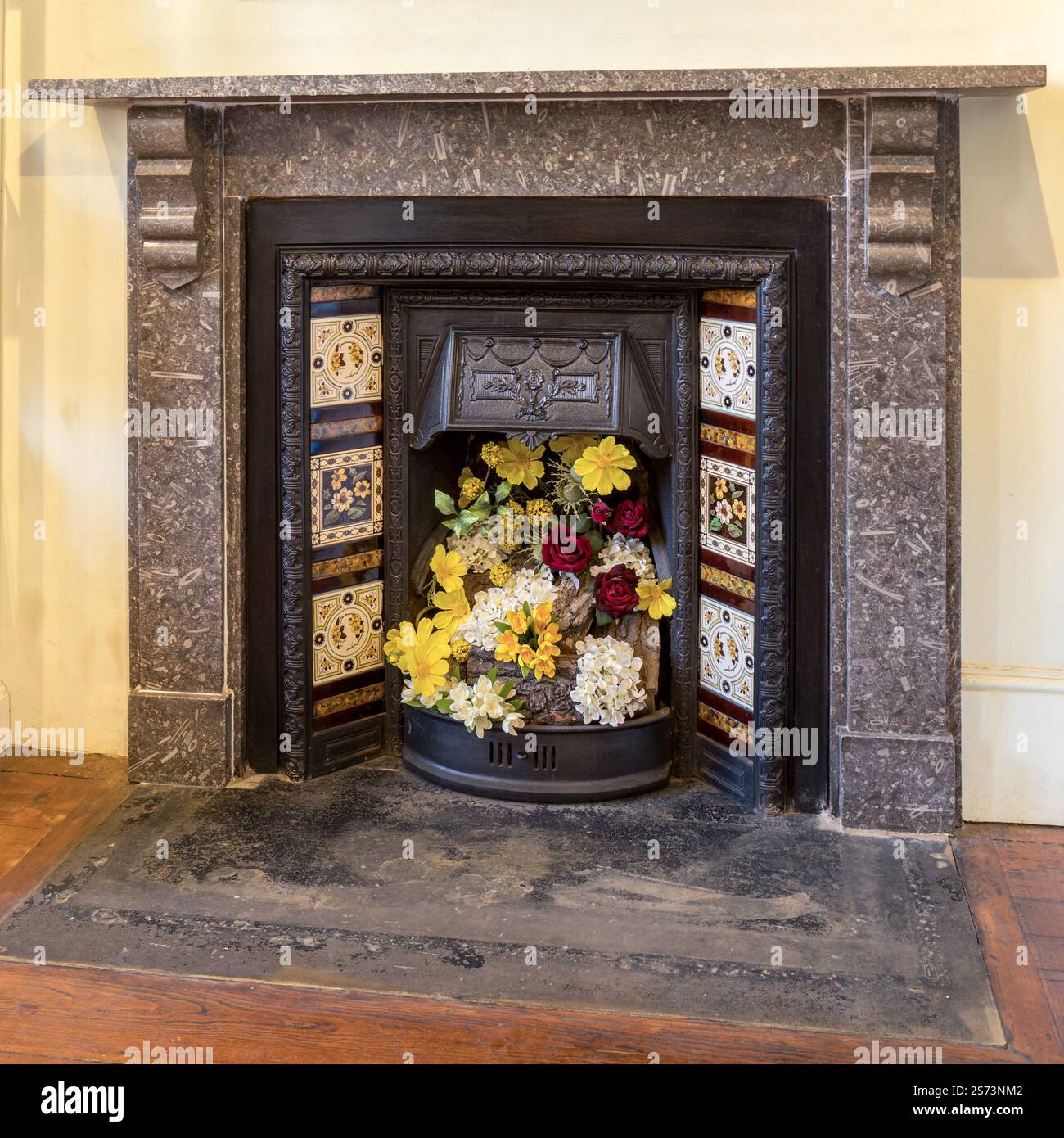 Cheminée victorienne en pierre calcaire fossile entourée d'une cheminée en fonte noire ornée d'ornements et d'une grille avec carreaux de céramique décoratifs et présentation de fleurs. Banque D'Images