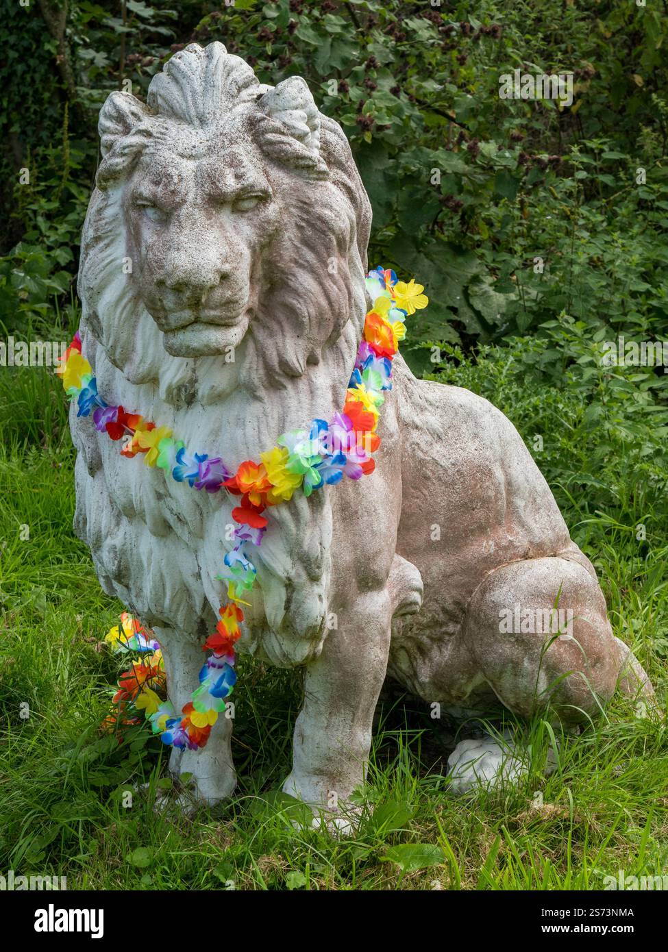 Grande statue ornementale de lion blanc décorée d'une guirlande de fleurs / collier autour de son cou dans le jardin domestique avec feuillage vert derrière, Royaume-Uni Banque D'Images
