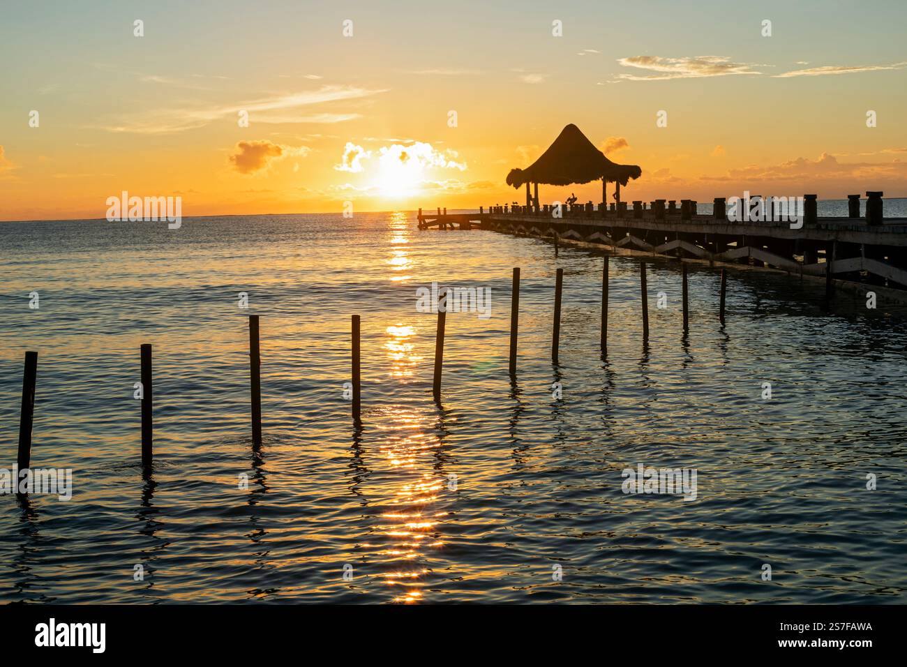Mexique, Puerto Morelos, jetée avec palapa et palmier au premier plan. Jeune femme en maillot de bain au bord de l'eau réjouissant l'eau. au lever du soleil Banque D'Images