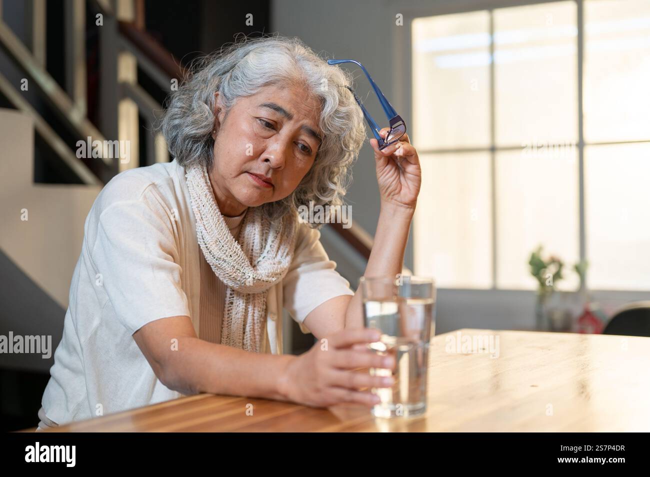 Une triste femme asiatique retraitée senior est assise sur un îlot de cuisine, regardant un verre d'eau avec un sentiment désespéré, se sentant seule après la retraite. Banque D'Images
