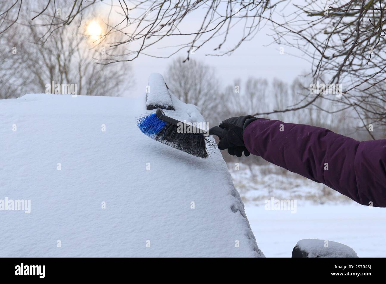 La main de la femme avec un balai à neige va nettoyer une épaisse couche de neige du pare-brise d'une voiture par une journée ensoleillée d'hiver Banque D'Images