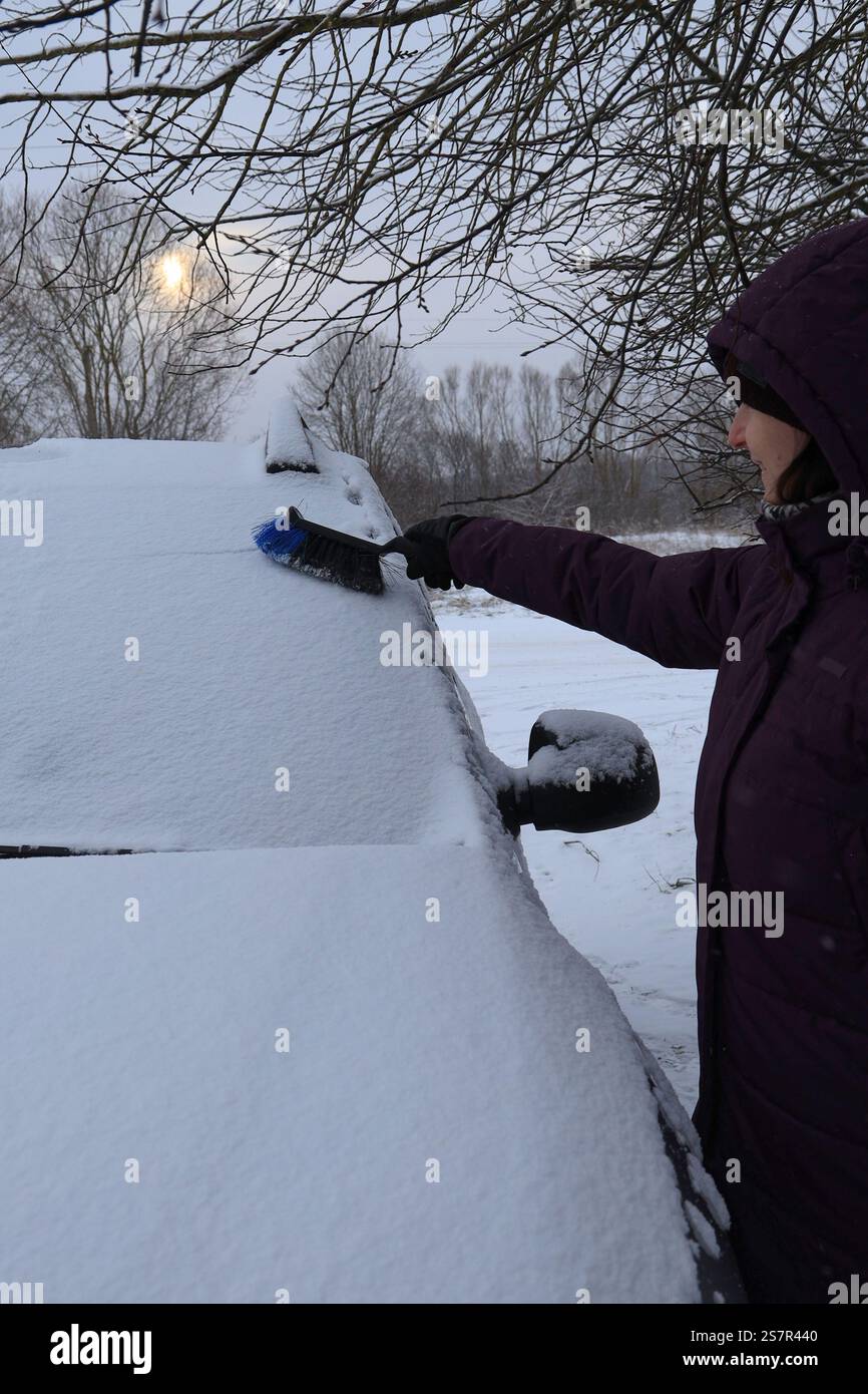 Jeune femme dans une veste chaude violette va nettoyer la neige du pare-brise d'une voiture avec un pinceau à neige Banque D'Images
