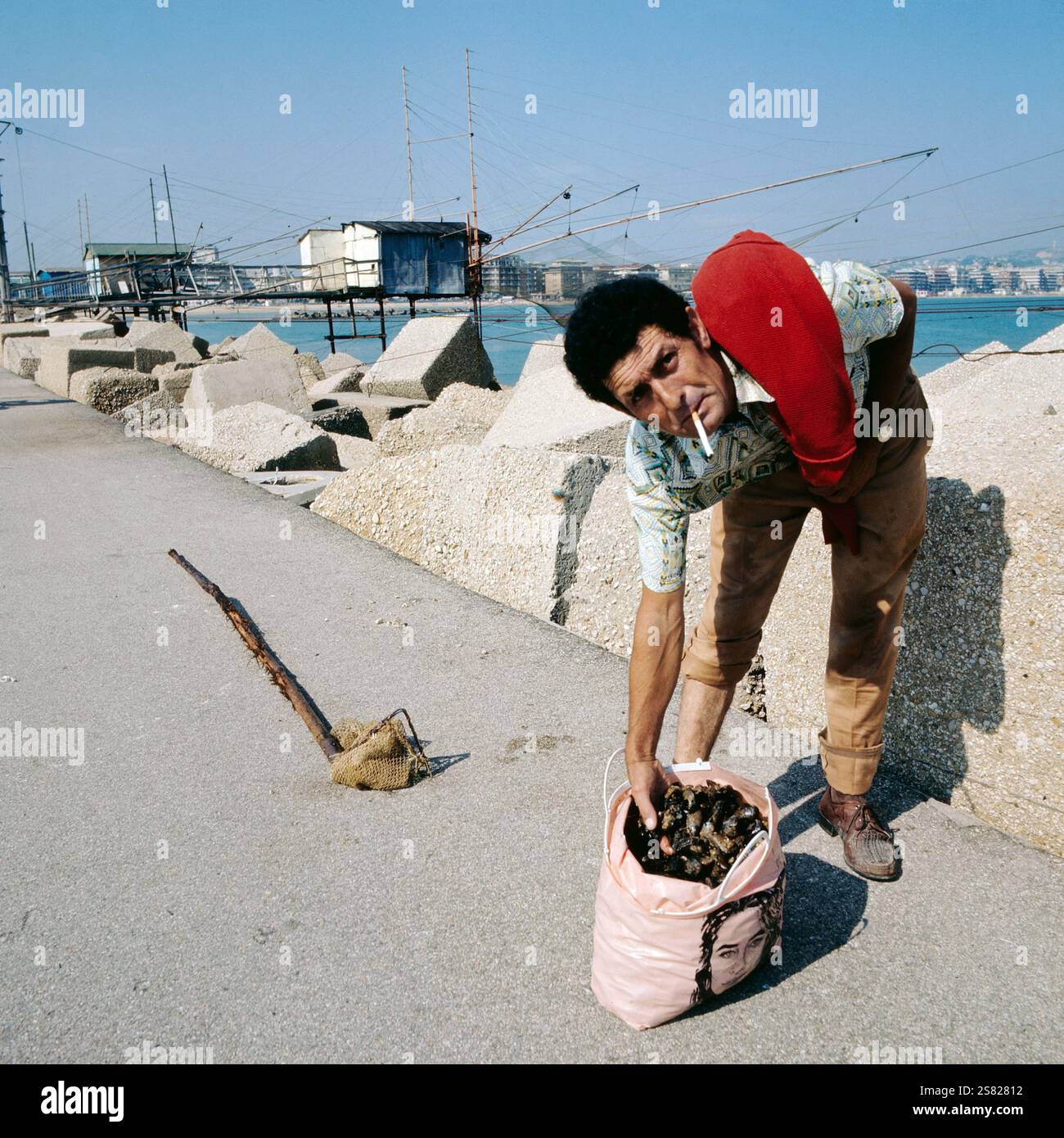 Italienischer Adria-Fischer hält an einem sonnigen Tag eine Tüte Muscheln in der Nähe der Fischernetze, Italien um 1977. Banque D'Images