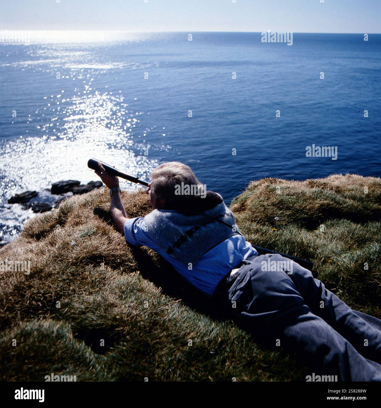 Ein Wissenschaftler beobachtet mit einem Teleskop von ihrem Aussichtspunkt auf einer grasbewachsenen Klippe aus, wie die Meereswellen auf die darunter liegenden Felsen krachen, Island um 1980. Banque D'Images