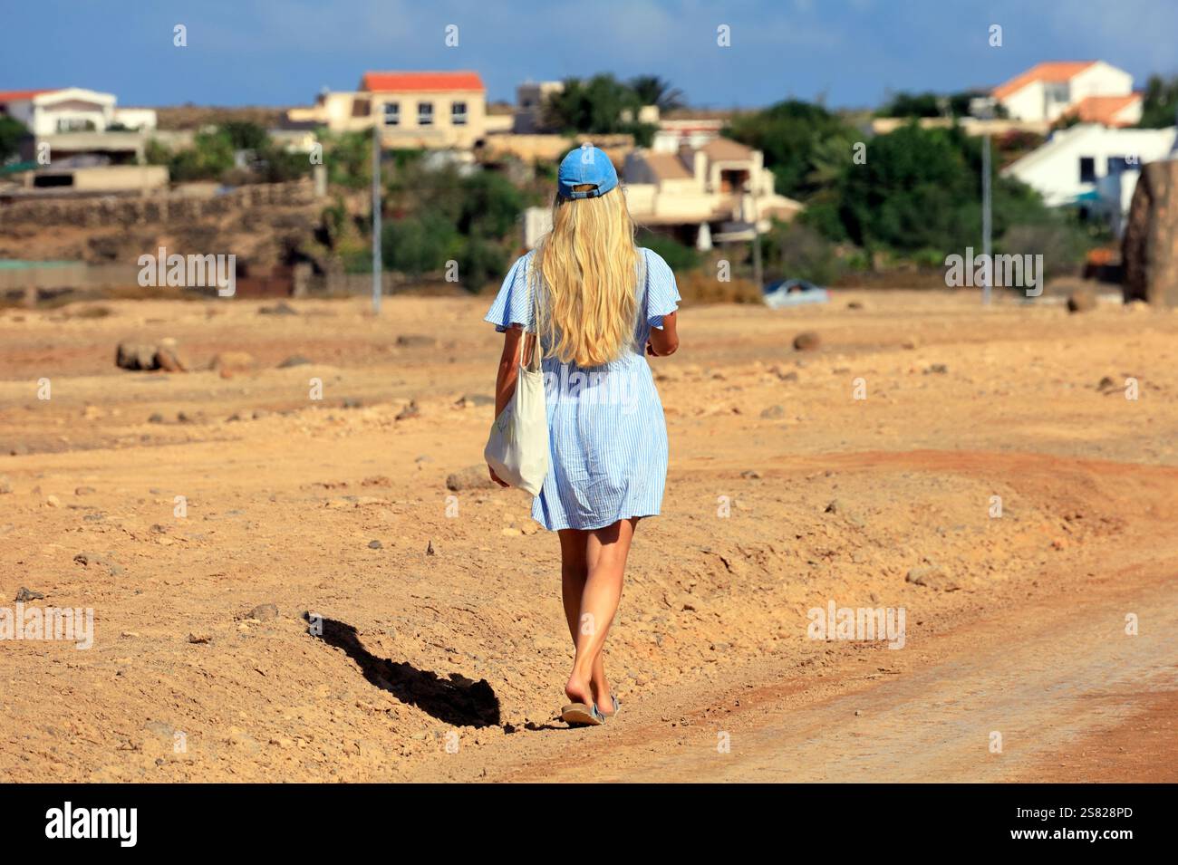 Femme blonde en robe bleue courte et chapeau marchant loin dans un chemin poussiéreux, Fuerteventura. Prise en décembre 2024. Banque D'Images