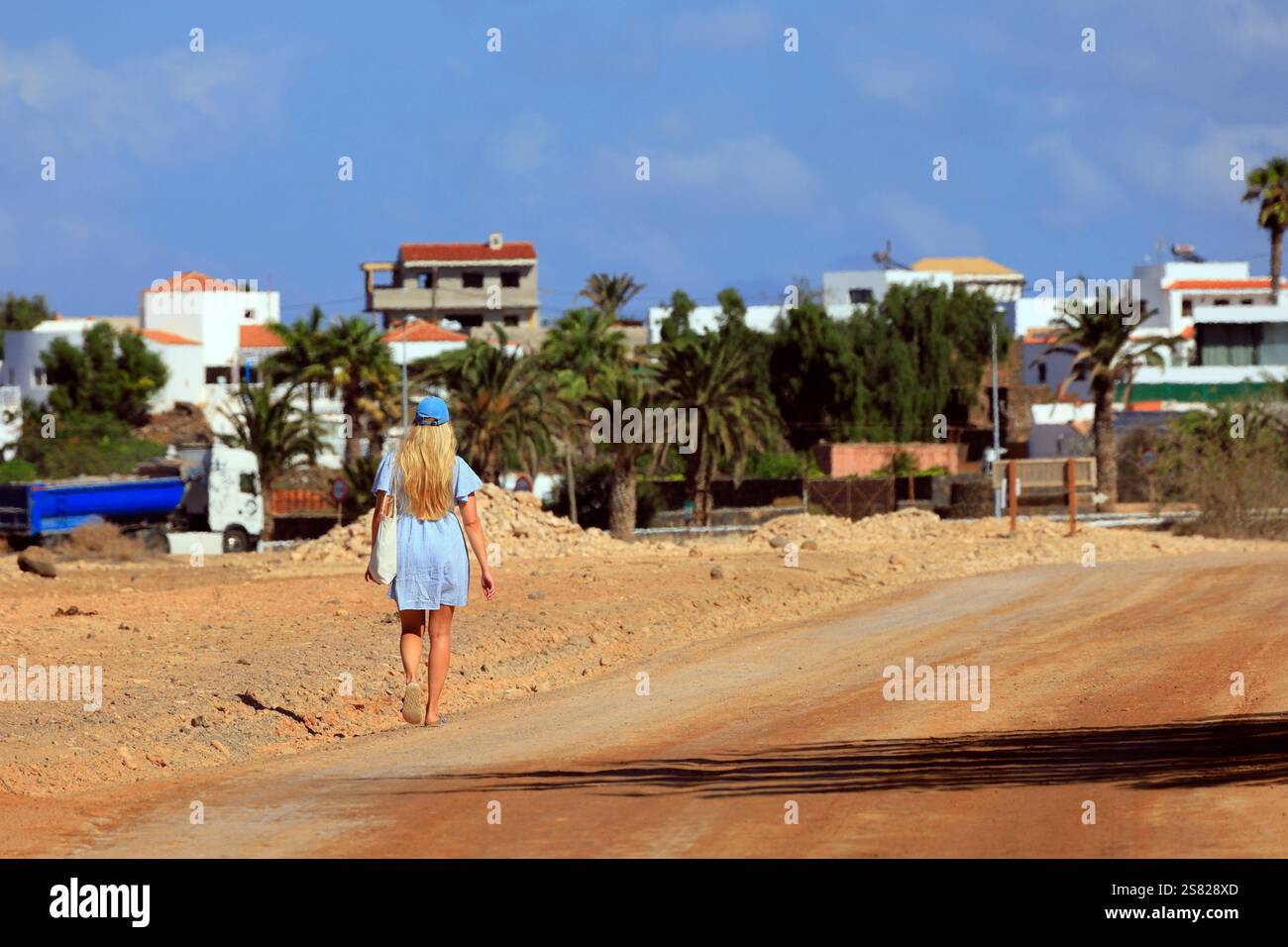 Femme blonde en robe bleue courte et chapeau marchant loin dans un chemin poussiéreux, Fuerteventura. Prise en décembre 2024. Banque D'Images