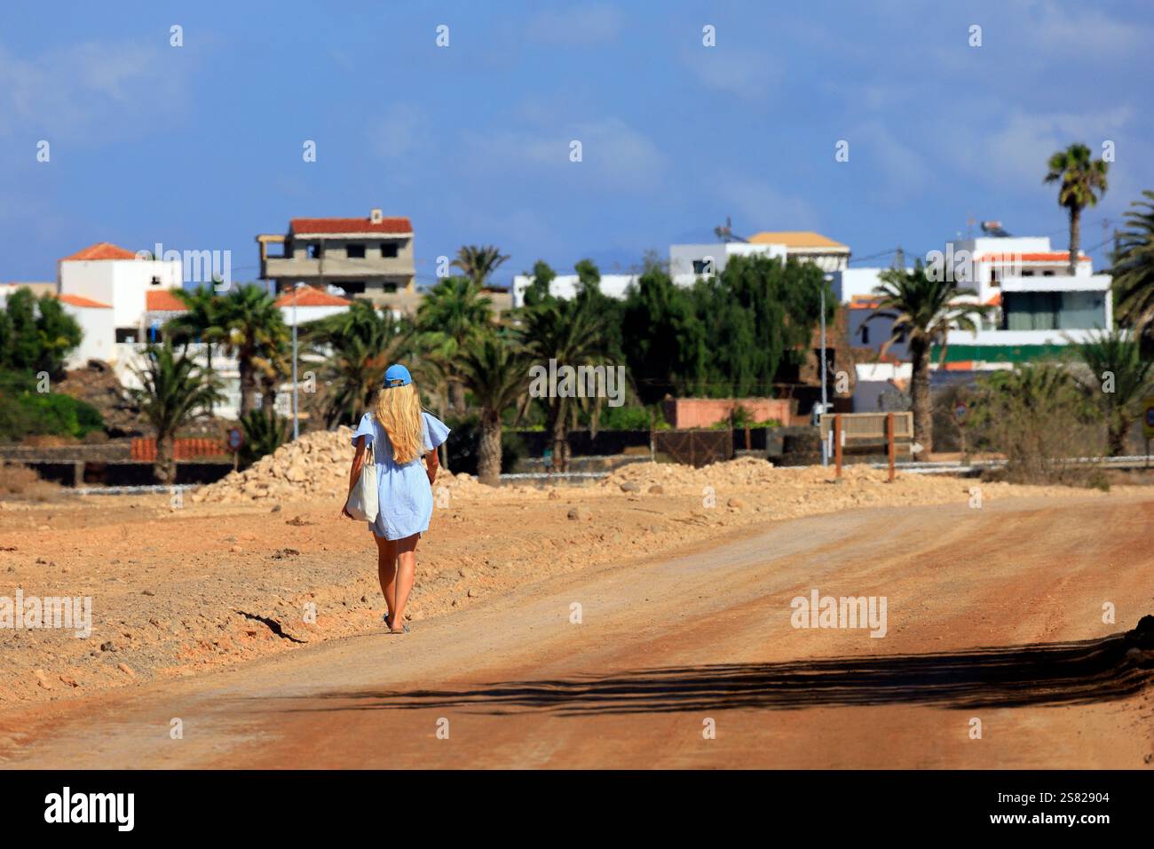 Femme blonde en robe bleue courte et chapeau marchant loin dans un chemin poussiéreux, Fuerteventura. Prise en décembre 2024. Banque D'Images
