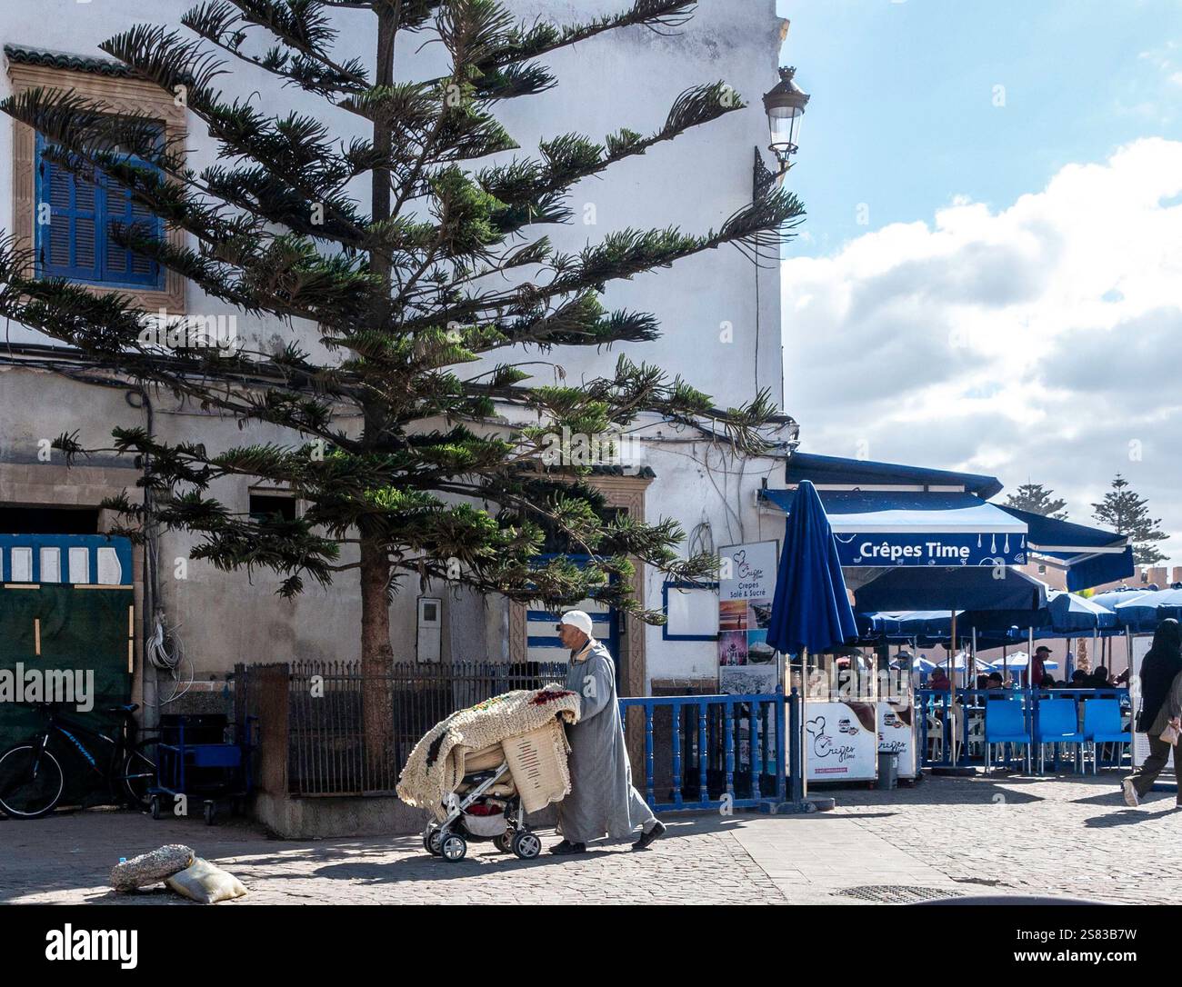 Un vendeur pousse une poussette chargée de tapis colorés dans les rues animées d'Essaouira, au Maroc, capturant l'artisanat local. Banque D'Images