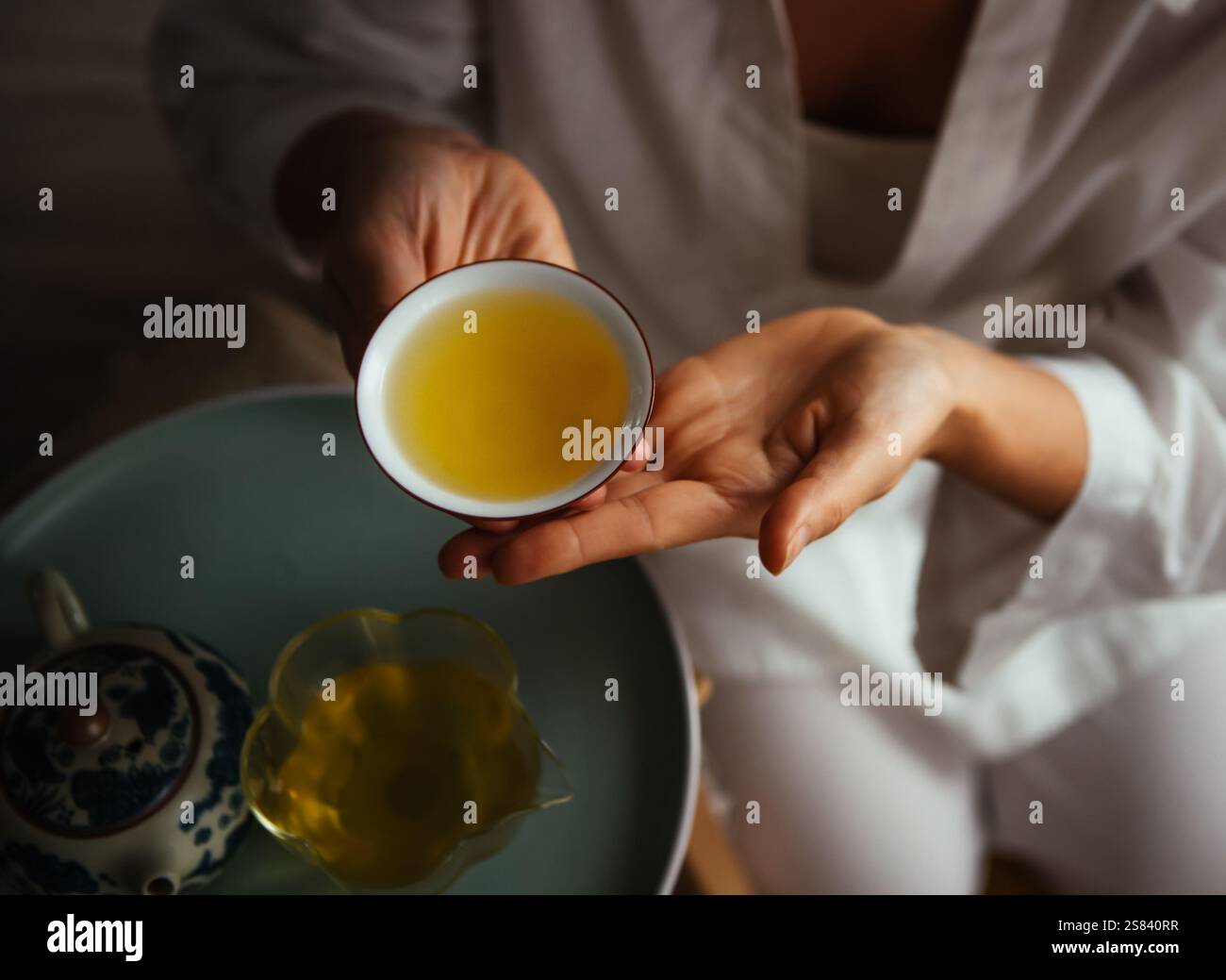 Mains tenant une tasse de porcelaine remplie de thé doré, exsudant chaleur et tranquillité. La configuration élégante avec théière et pichet en verre renforce l'intimité Banque D'Images