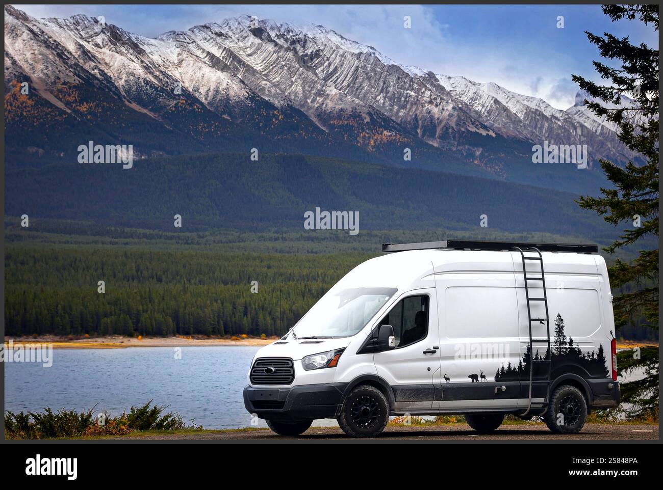 Un camping-car garé au bord d'un lac tranquille, entouré de forêts luxuriantes et de montagnes enneigées, présentant l'escapade d'aventure parfaite. Banque D'Images