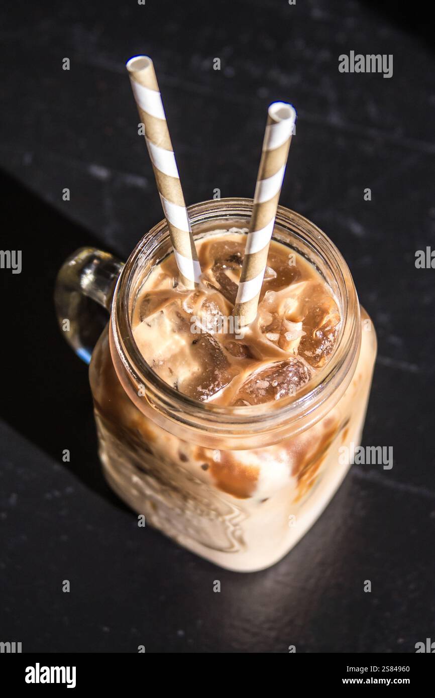 Un pot mason en verre rempli de café glacé et de glaçons repose sur une surface sombre. La lumière du soleil crée des ombres nettes, accentuant le contraste de la boisson. Banque D'Images