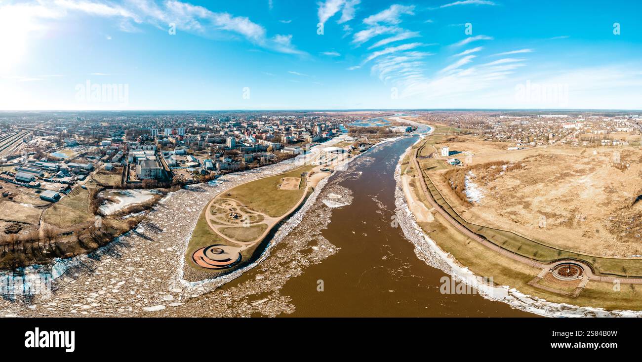 Vue aérienne panoramique d'une ville divisée par une rivière partiellement gelée, avec des bâtiments urbains, des champs ouverts et une structure en spirale circulaire près du Banque D'Images